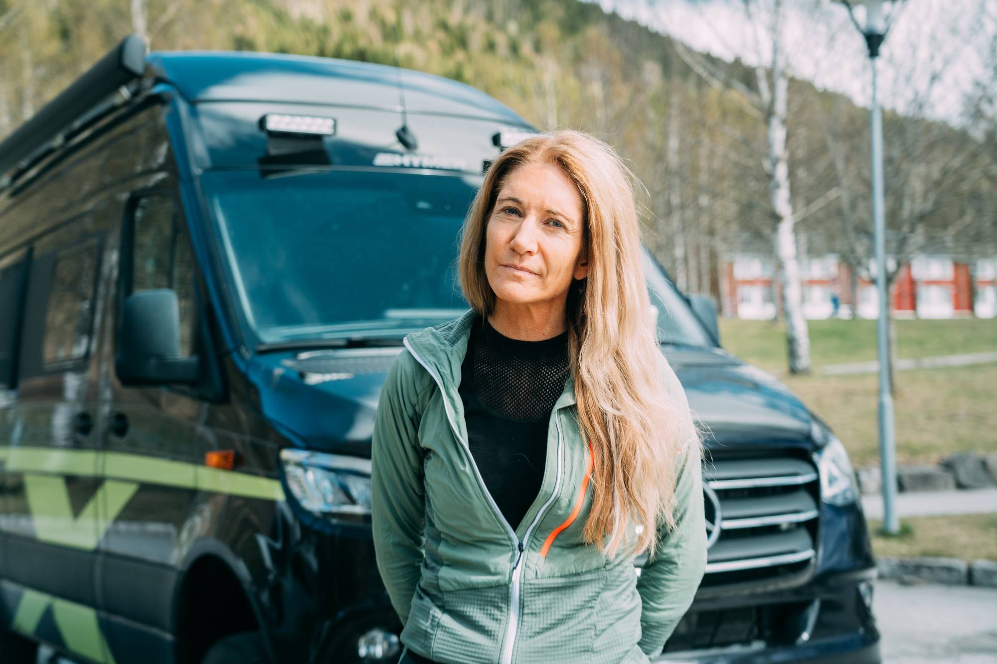 En kvinne står foran en mørk varebil. Hun har blondt hår og har på seg en grønn jakke over en svart topp. Varebilen har gule striper langs siden og er parkert utendørs. I bakgrunnen er det trær og røde bygninger. (Bildebeskrivelsen er laget av en KI-tjeneste)