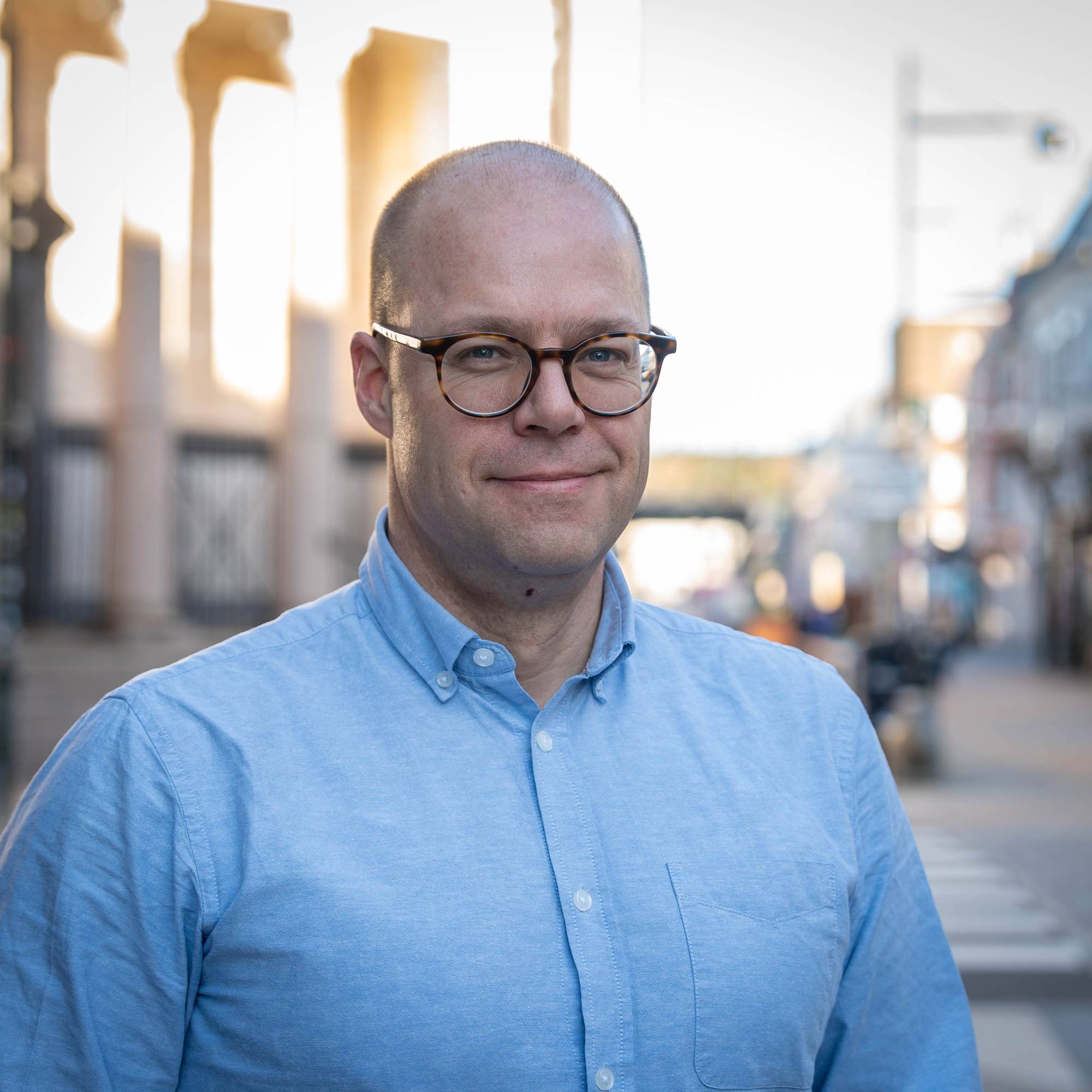 En mann med briller og lyseblå skjorte ser mot kameraet. I bakgrunnen er en bygate med bygninger og en gangovergang. Sollyset er sterkt i bakgrunnen. (Bildebeskrivelsen er laget av en KI-tjeneste)