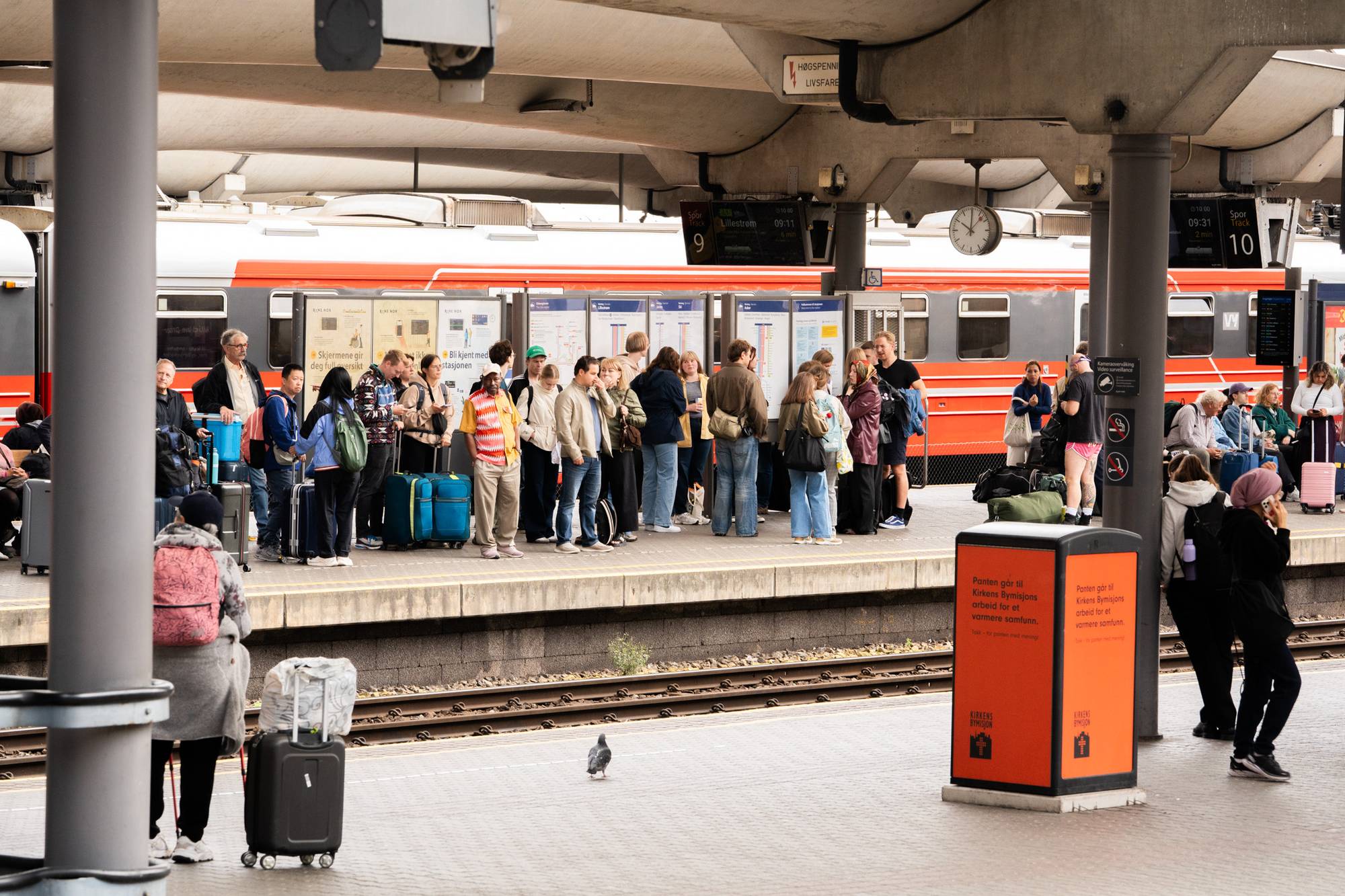 Mennesker venter på tog på en perrong. Toget står parkert ved sporene i bakgrunnen. 