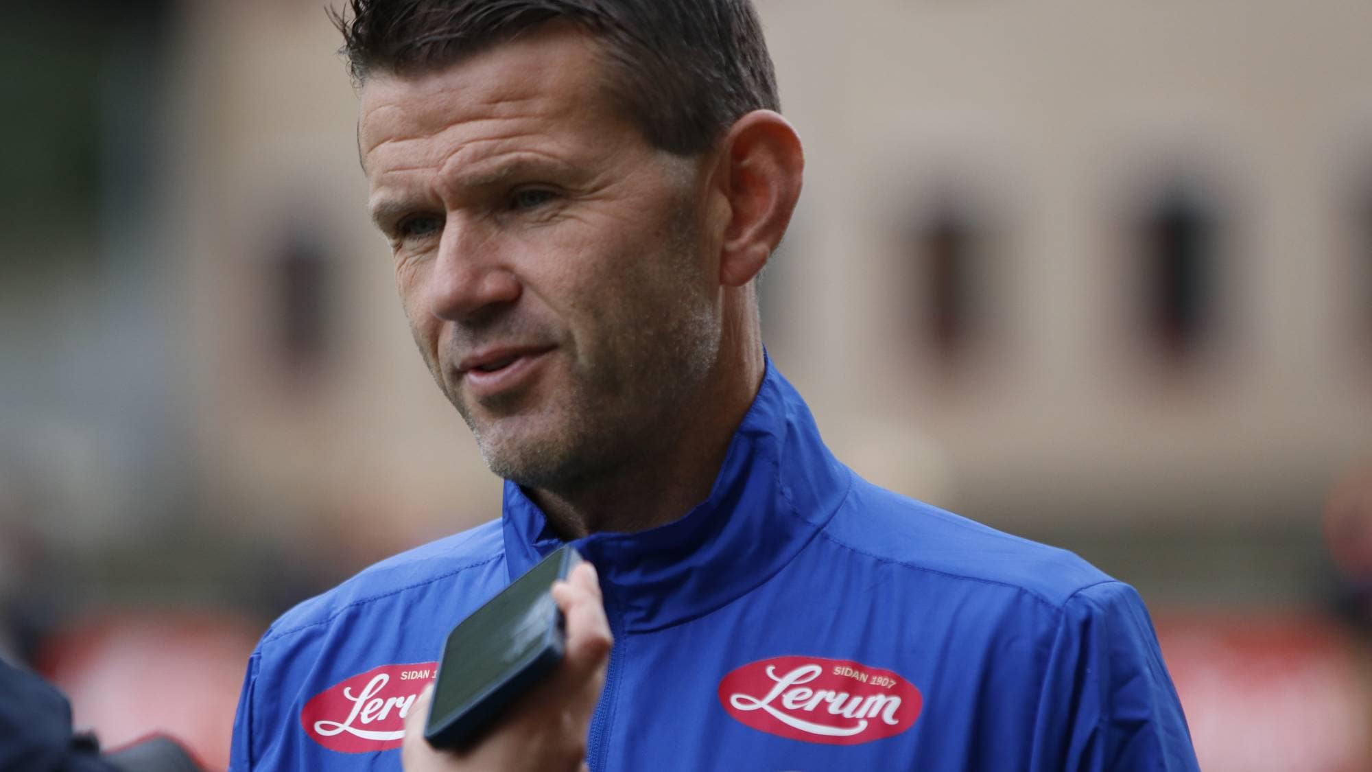 Fotballtrener Eirik Bakke intervjues og blir holdt opp en mobiltelefon for lydopptak. (Bildebeskrivelsen er laget av en KI-tjeneste)