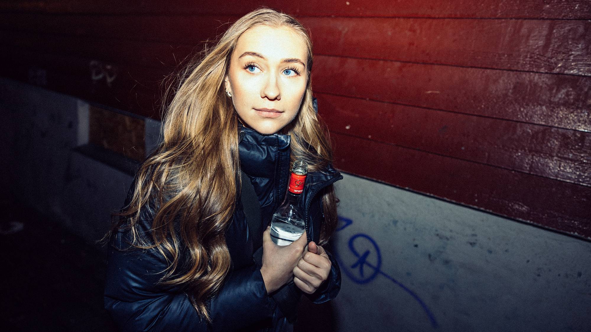 Julie (23) har langt hår med lette krøller og store blå øyne. Hun skjuler en vodkaflaske med vann i den mørke boblejakken sin. Hun ser til venstre med et litt urolig blikk mens hun går forbi en vegg med graffiti i et smug.