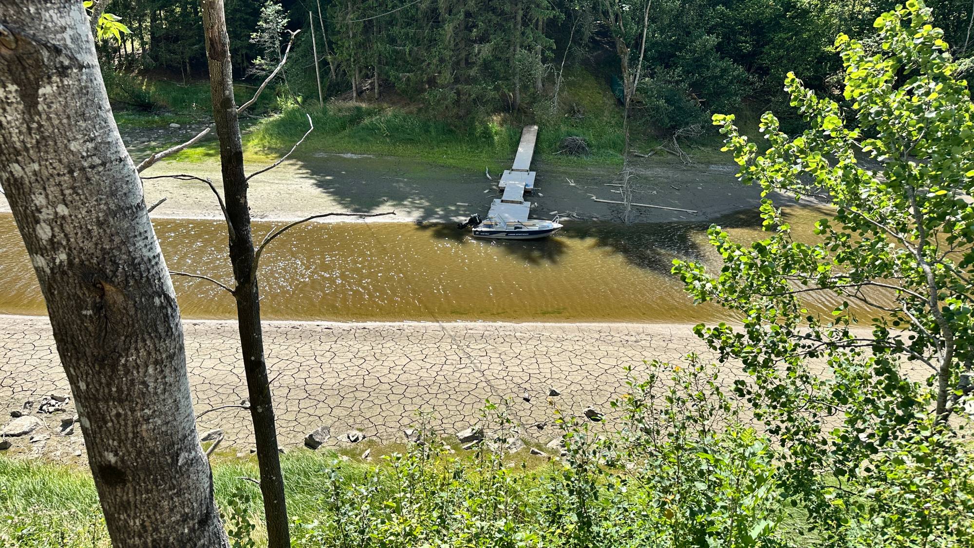 En liten båt ligger fortøyd ved en brygge i en uttørket innsjø med sprukken mudderbunn. (Bildebeskrivelsen er laget av en KI-tjeneste)