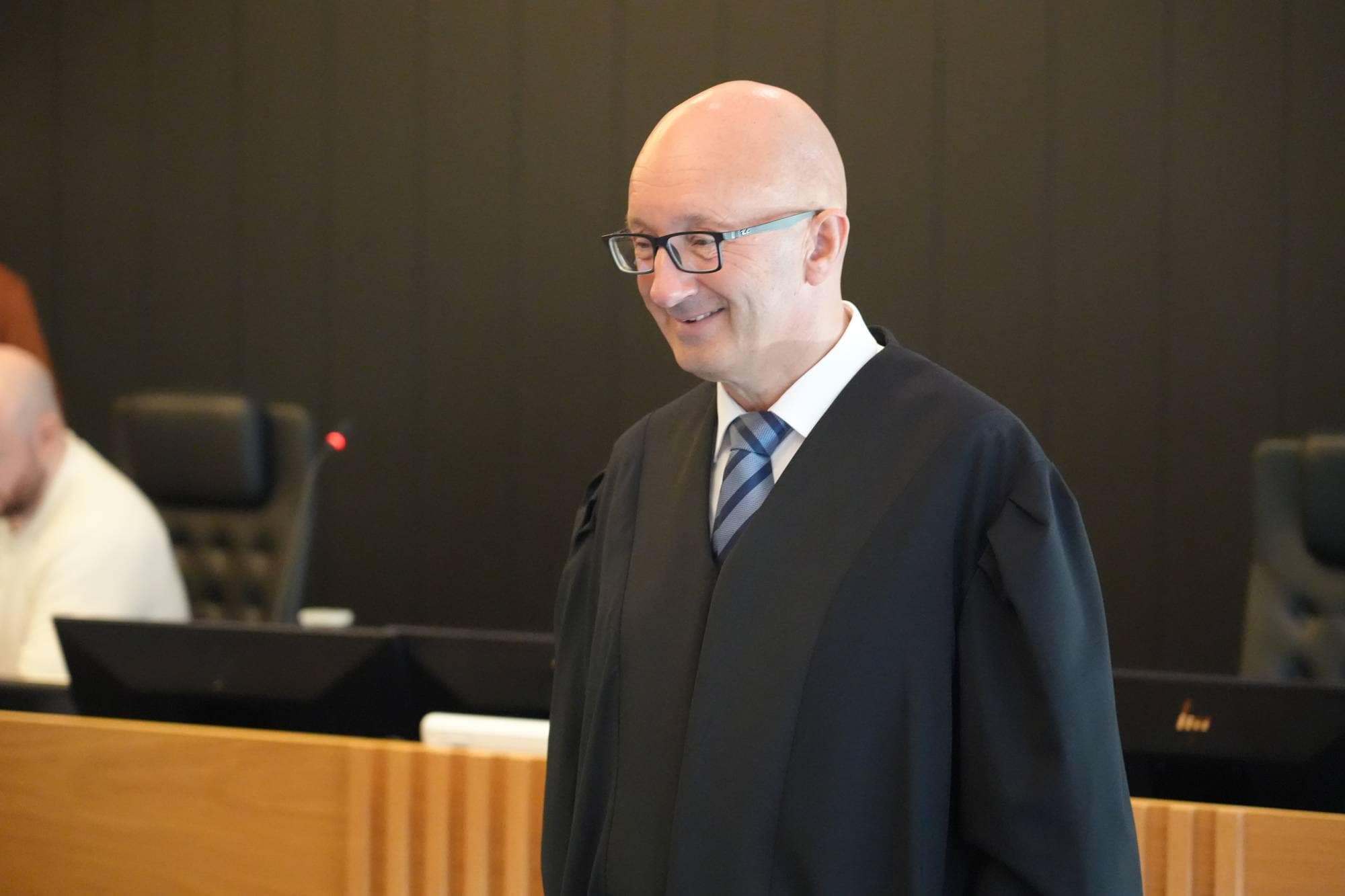 Skallet mann med briller i dommerkappe står i en rettssal.
