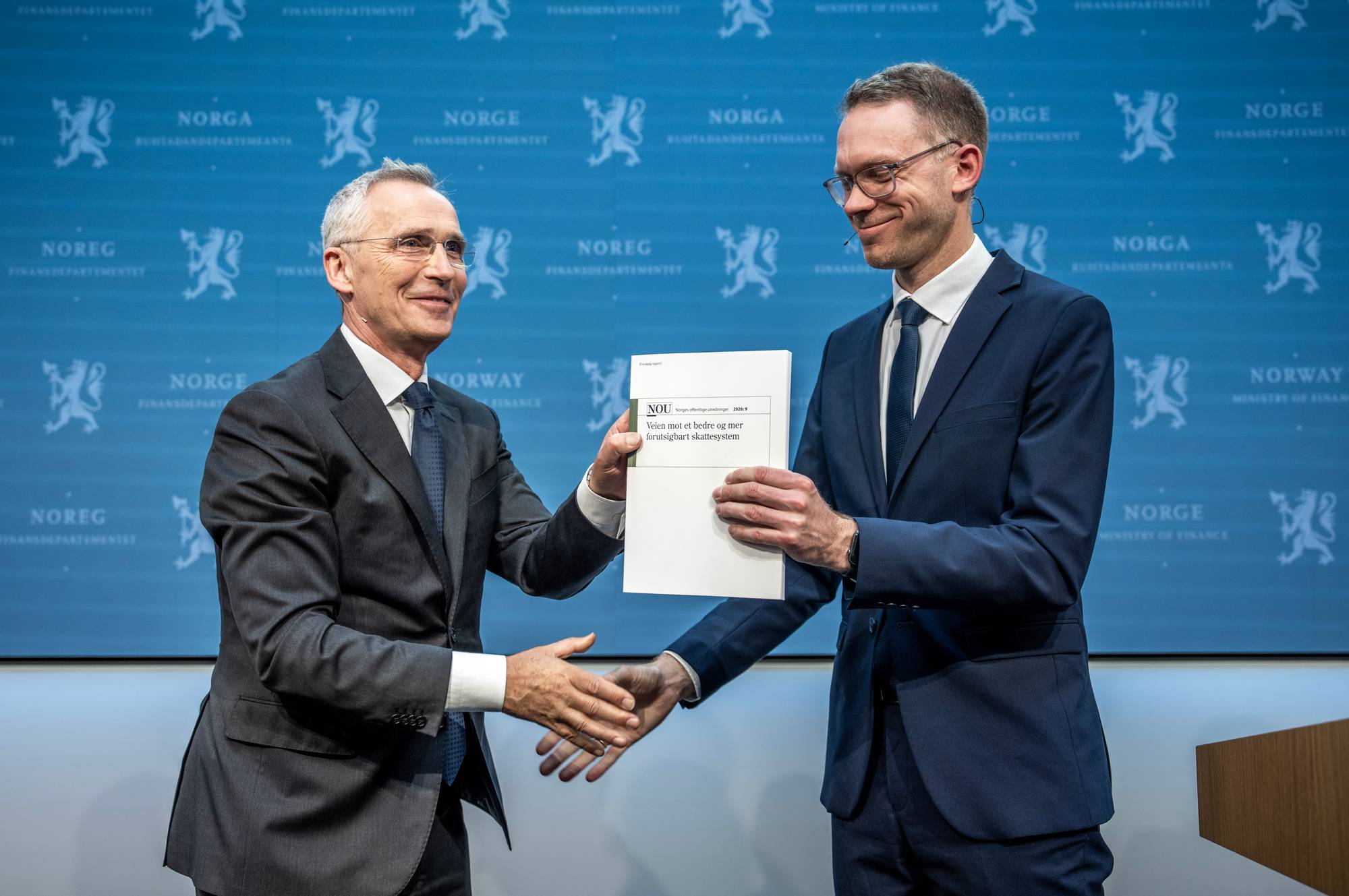 To menn i dress gir et dokument til hverandre. Mannen til venstre overrekker dokumentet til mannen til høyre, som tar imot. Bak dem er et blått bakteppe med gjentatte symboler og tekst om Norges Finansdepartement. (Bildebeskrivelsen er laget av en KI-tjeneste)