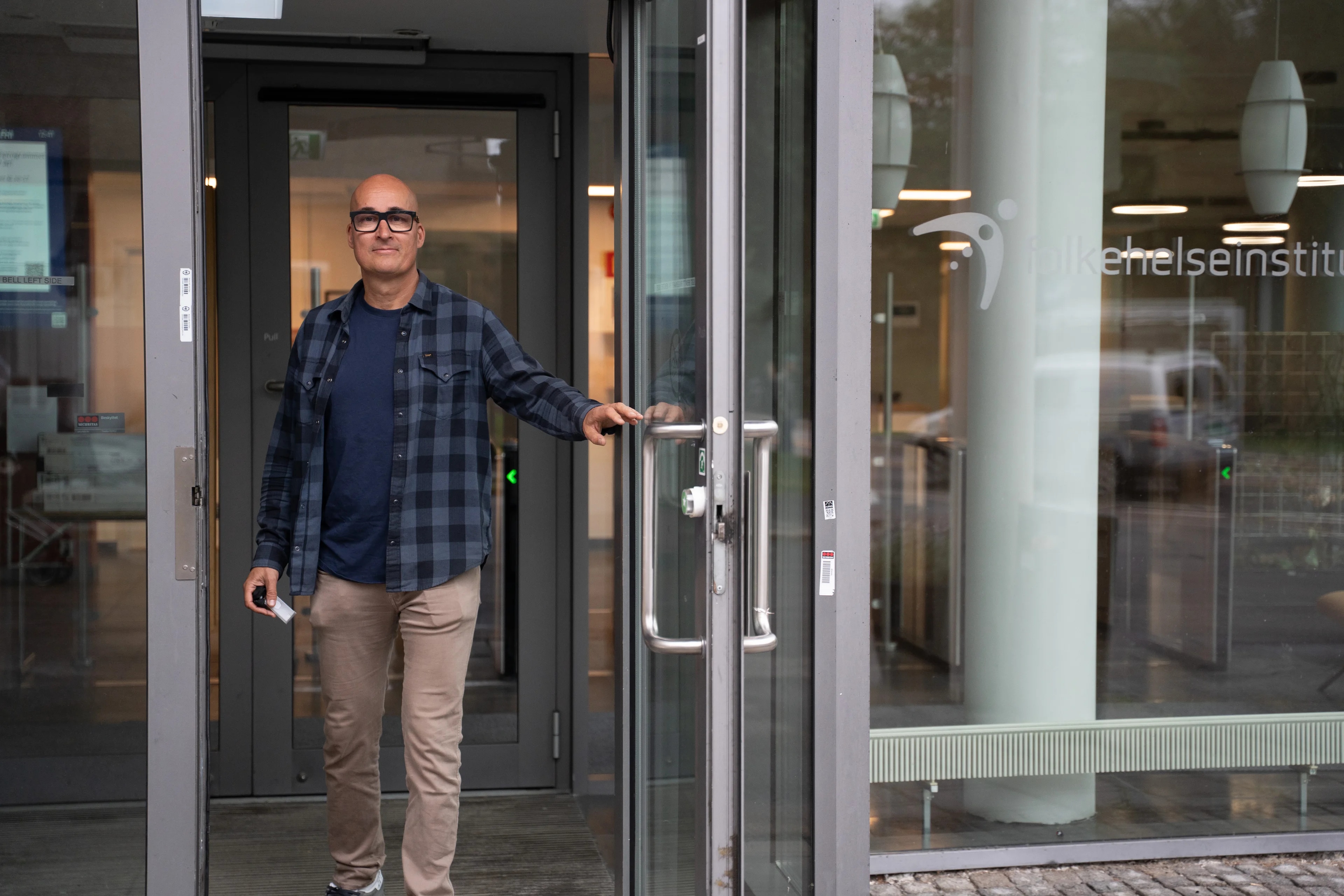 Skallet mann i rutete skjorte åpner glassdøren til en bygning. (Bildebeskrivelsen er laget av en KI-tjeneste)