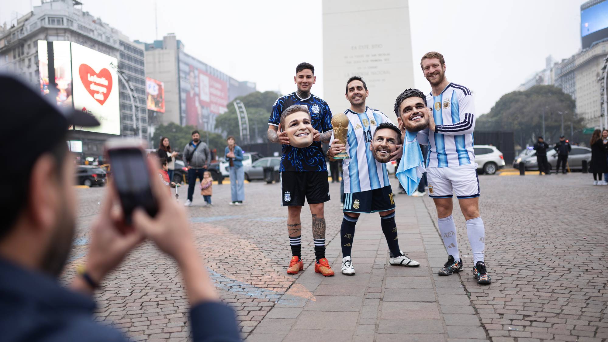 Tre menn i fotballdrakter poserer med masker av landslagsspillere foran Obelisken i Buenos Aires.
