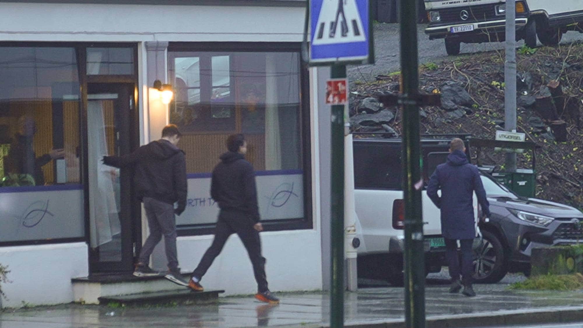 Tre personer, iført mørke klær, forlater en bygning med vinduer mot gaten. En av dem holder døren åpen for de andre mens de går ned en trapp. I bakgrunnen står to biler parkert langs veien.