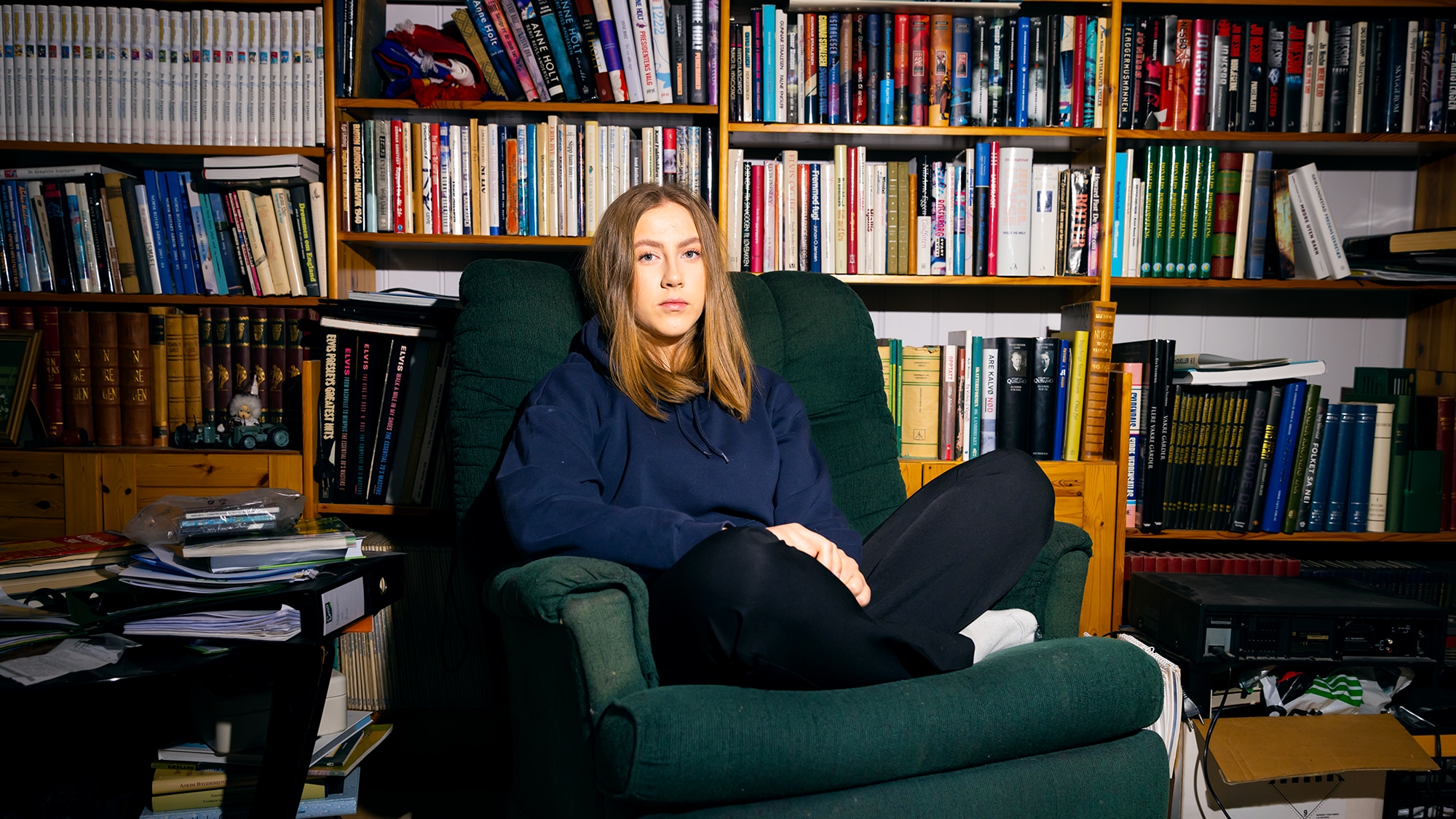 Andrea (17) sitter med beina i kryss i en stor lenestol foran masse bøker. Hun har midtskill hettegenser dressbukse og et et seriøst blikk.