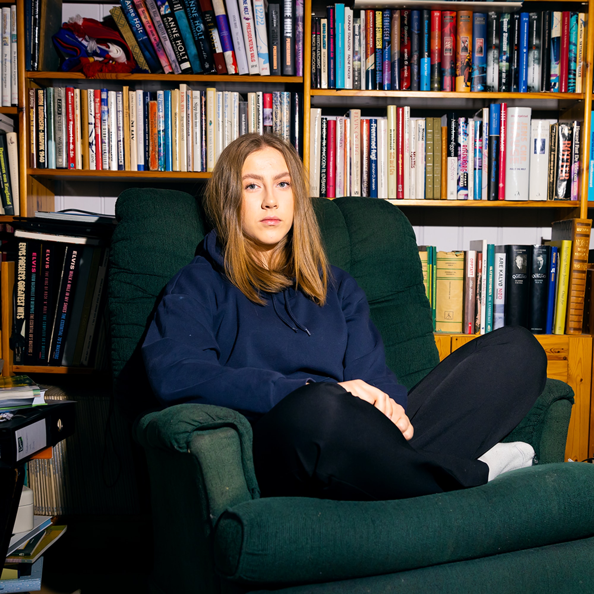 Andrea (17) sitter med beina i kryss i en stor lenestol foran masse bøker. Hun har midtskill hettegenser dressbukse og et et seriøst blikk.