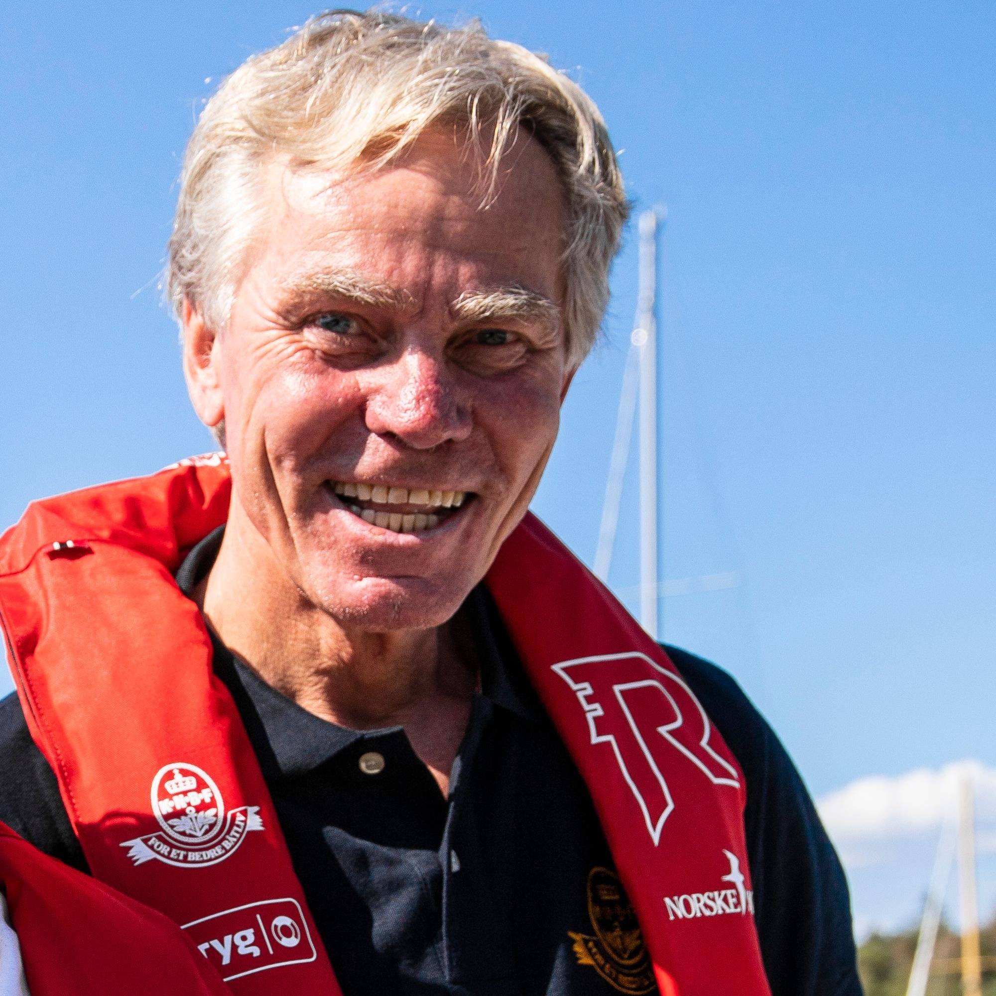 En person iført en rød flytevest står på en havn. Bak personen er seilbåter og blå himmel. Personen ser mot kameraet, og det er tydelig solskinn. Det er ingen andre synlige personer i nærheten.