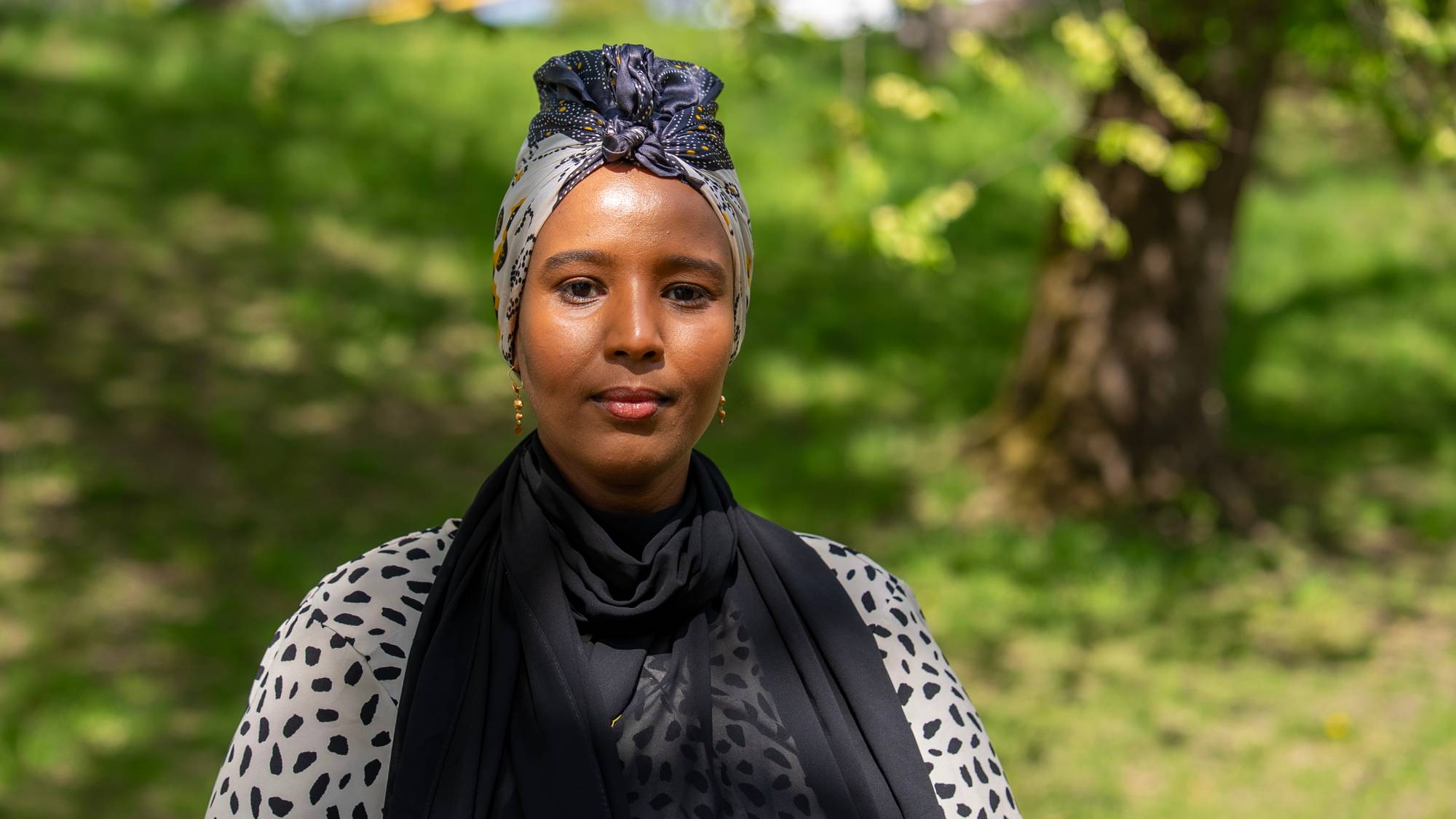 En kvinne med hijab står ute i sollys. Kvinnen har på seg en sort og hvit prikkete bluse med en sort sjal rundt halsen. Bak henne er det et tre og grønt gress. (Bildebeskrivelsen er laget av en KI-tjeneste)