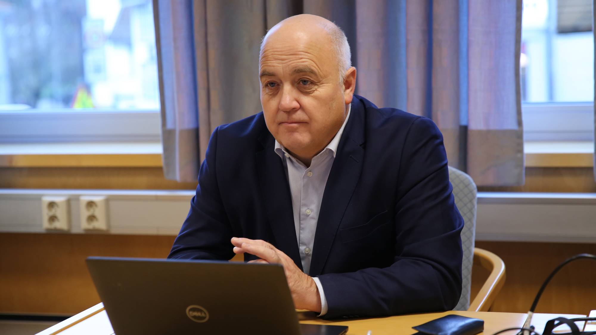 Mann kledd i dress sitter ved et bord med en laptop foran seg.