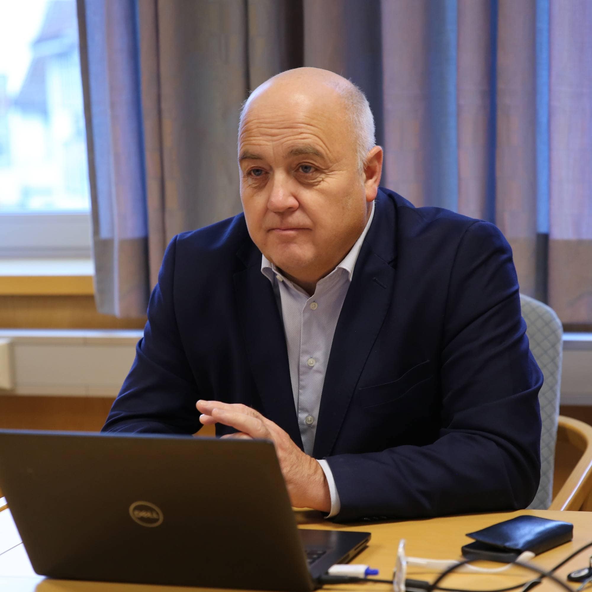 Mann kledd i dress sitter ved et bord med en laptop foran seg.