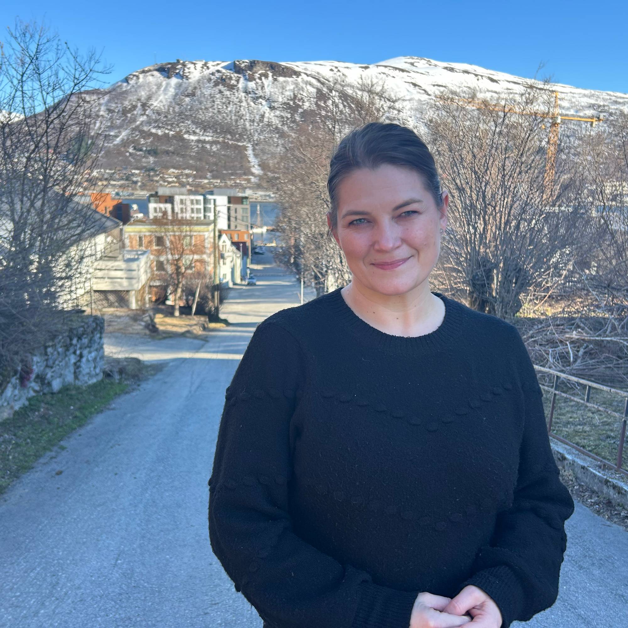 Kvinner i svart genser og mørkt oppsett hår står mot kamera, fjell i bakgrunnen.