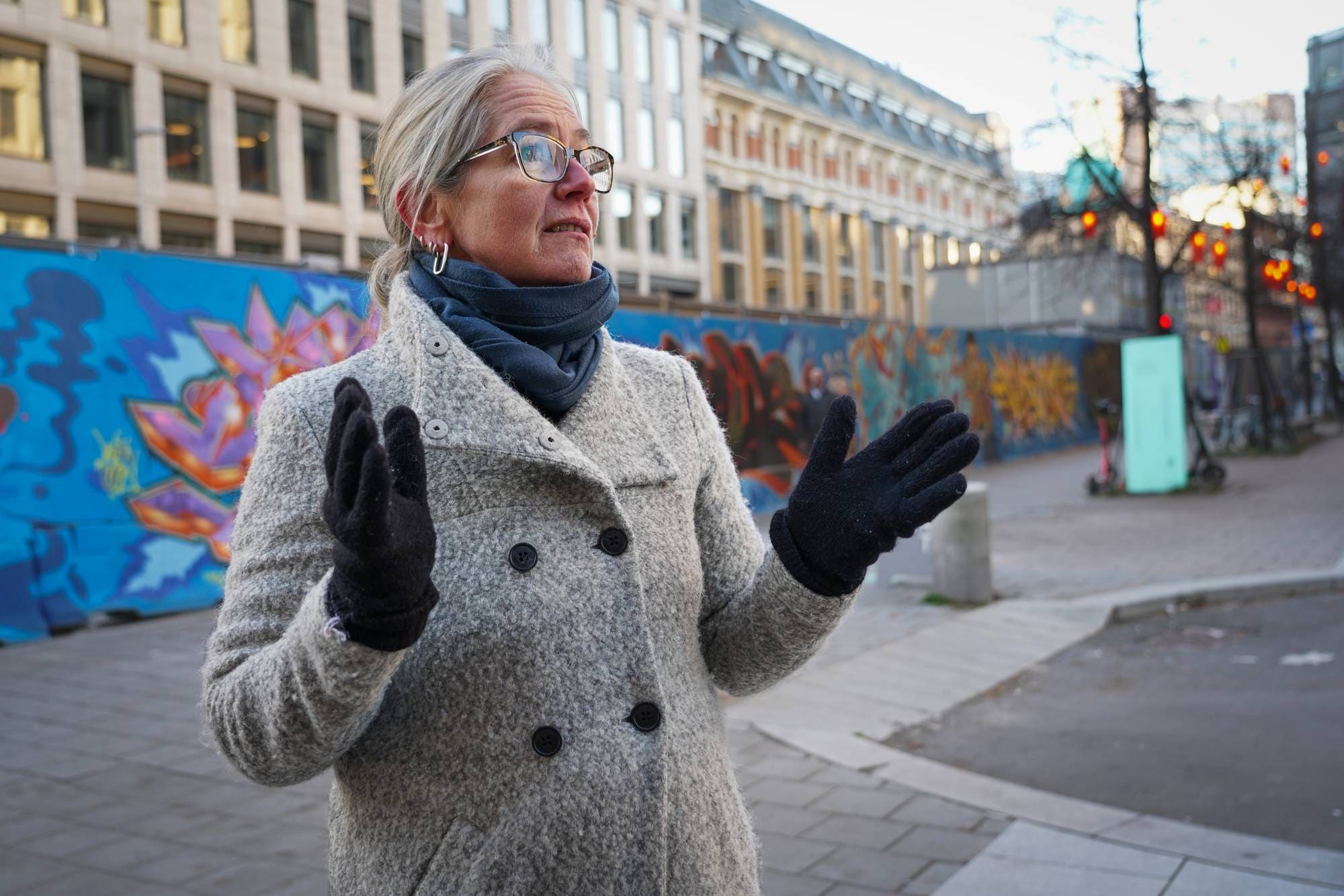 En person i grå frakk og mørke hansker står utendørs og gestikulerer. Bakgrunnen viser en muralvegg med fargerik graffiti. Det er en klar himmel, og bygninger vises på begge sider. Området ser ut til å være urbant og livlig.