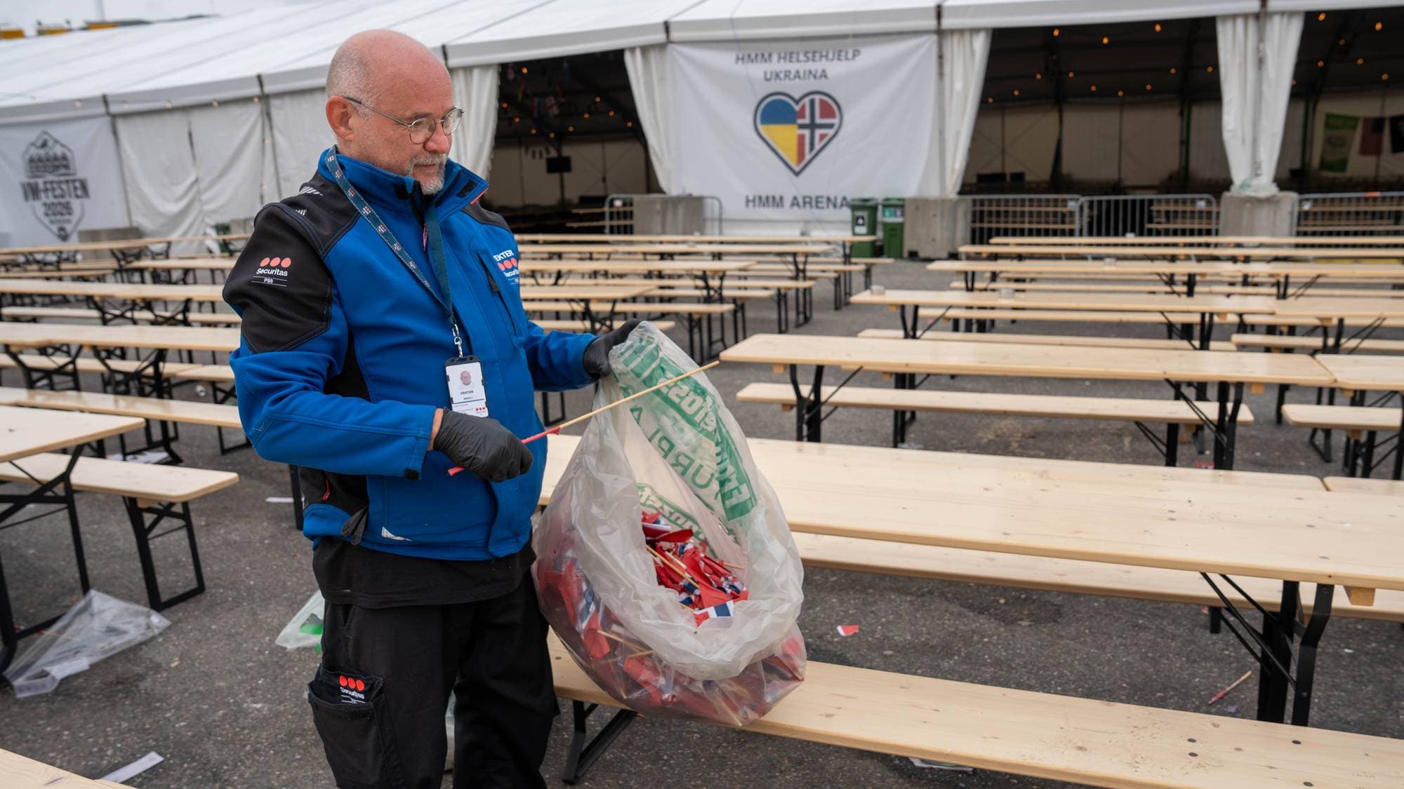 En mann i blå jakke og hansker holder en stor plastpose med norske flagg. Bak ham står mange tomme benker under et stort telt. Et banner med tekst og hjerteflagg henger på teltveggen. (Bildebeskrivelsen er laget av en KI-tjeneste)