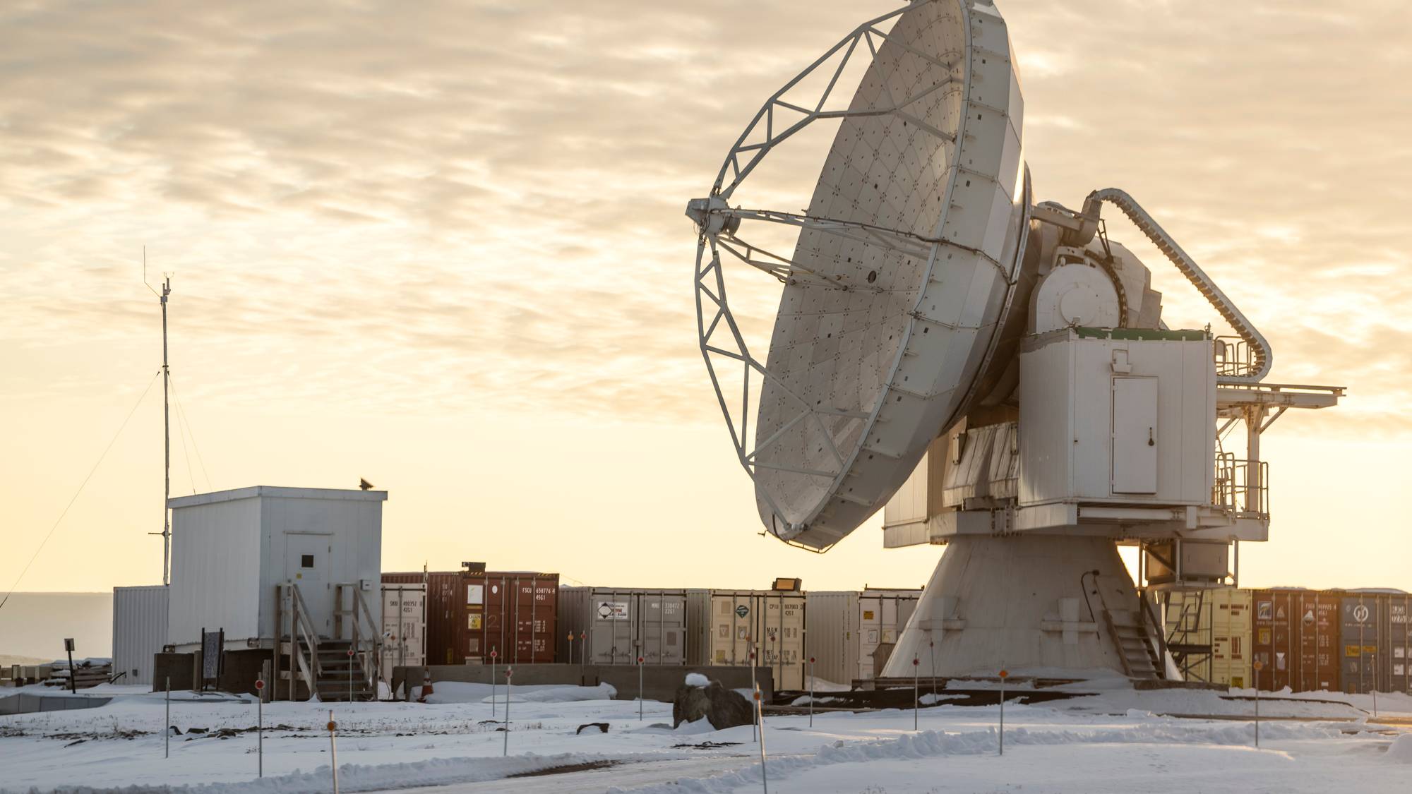 Stor parabolantenne plassert i et snødekket landskap med flere containere i bakgrunnen. (Bildebeskrivelsen er laget av en KI-tjeneste)