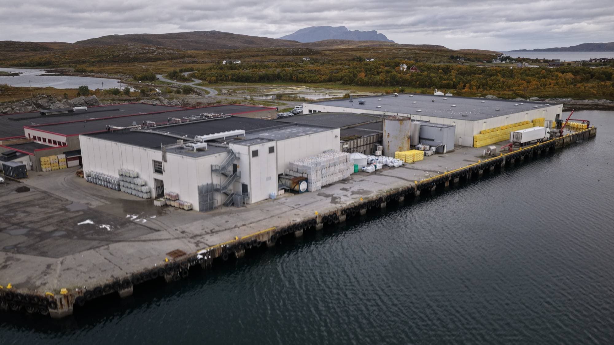 Industrianlegg ved kai med lagrede sekker og paller, sett ovenfra i et kystlandskap. (Bildebeskrivelsen er laget av en KI-tjeneste)