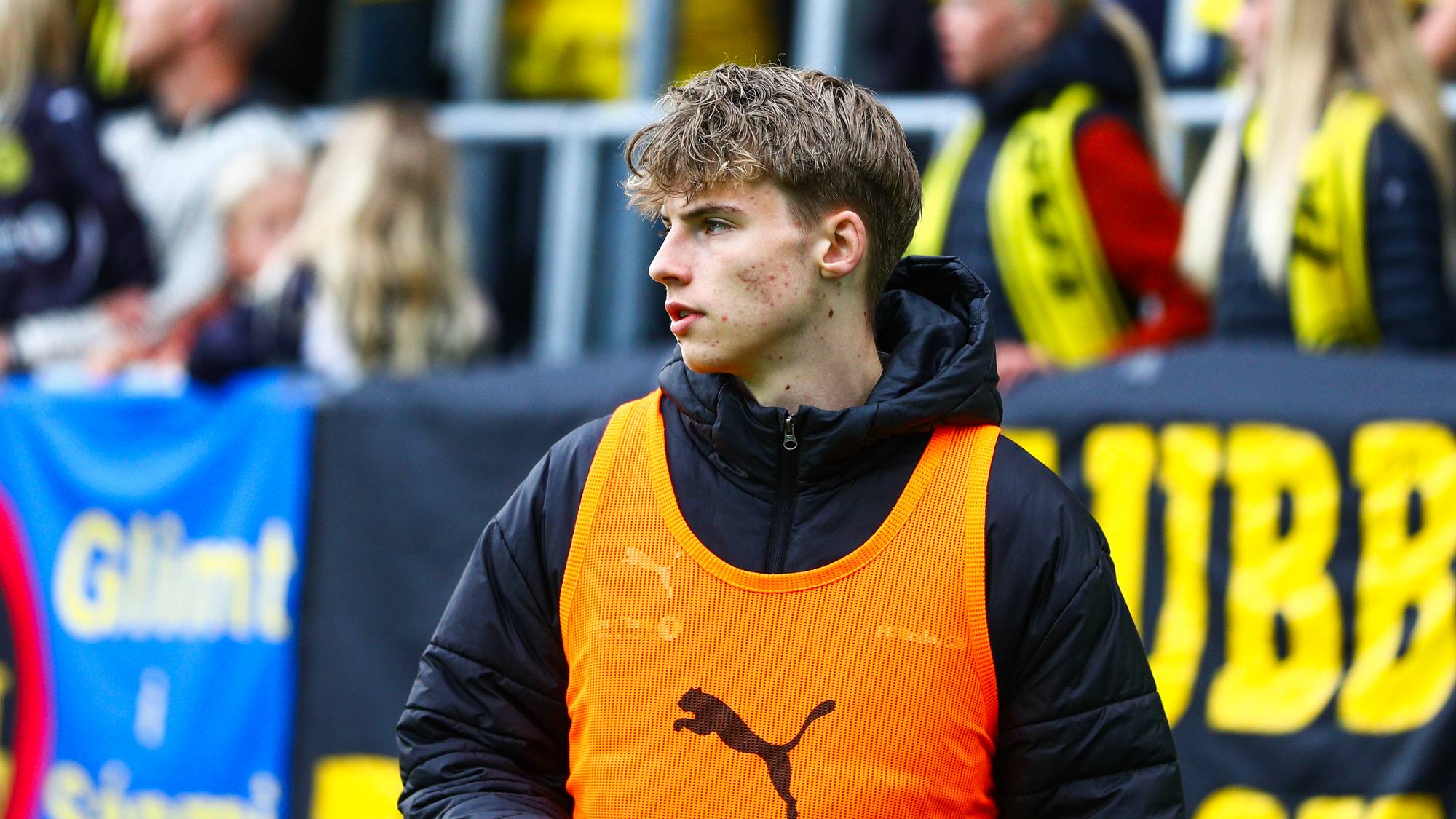 Fotballspiller med oransje vest på sidelinjen under kamp med supportere i bakgrunnen. (Bildebeskrivelsen er laget av en KI-tjeneste)