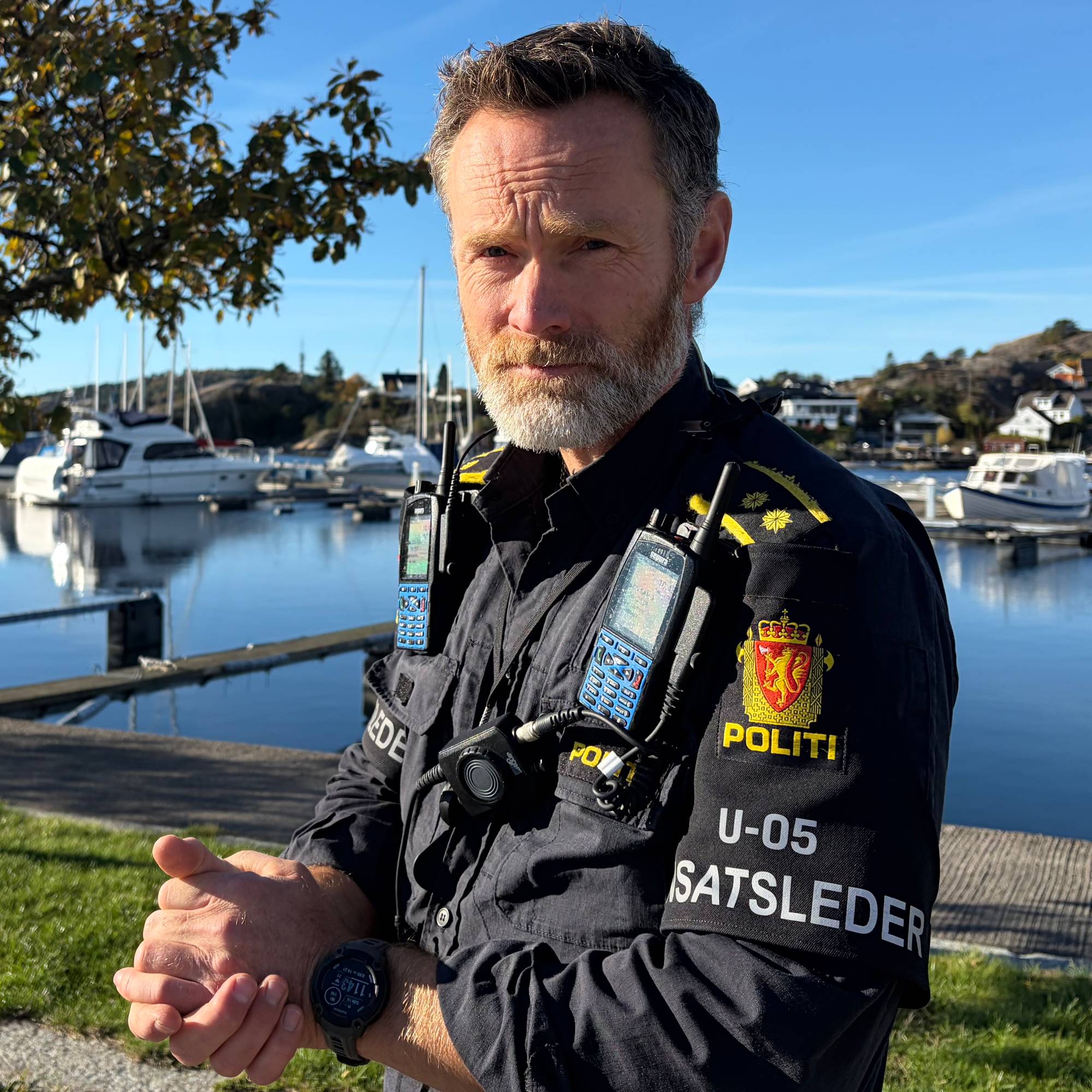 En politiinnsatsleder står ved en småbåthavn med fritidsbåter i bakgrunnen. (Bildebeskrivelsen er laget av en KI-tjeneste)