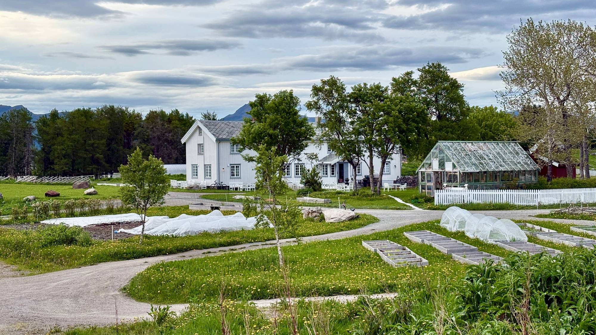Et hvitt hus med flere vinduer står i en grønn hage. En drivhus i glass er plassert til høyre for huset. I forgrunnen er det flere blomsterbed og en grusvei. (Bildebeskrivelsen er laget av en KI-tjeneste)