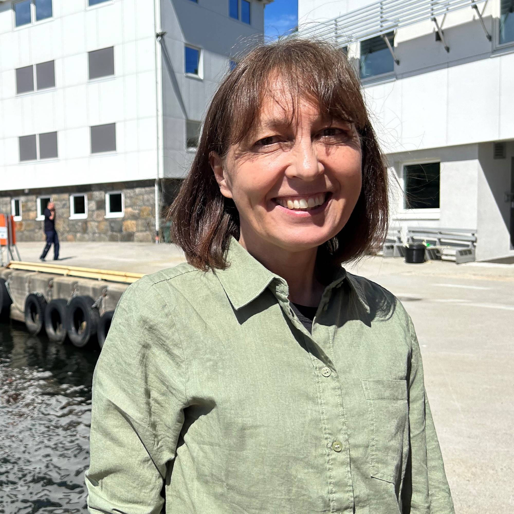 En kvinne smiler mot kameraet. Hun står ved en kai med sjøen til venstre og bygningsmasse til høyre. 