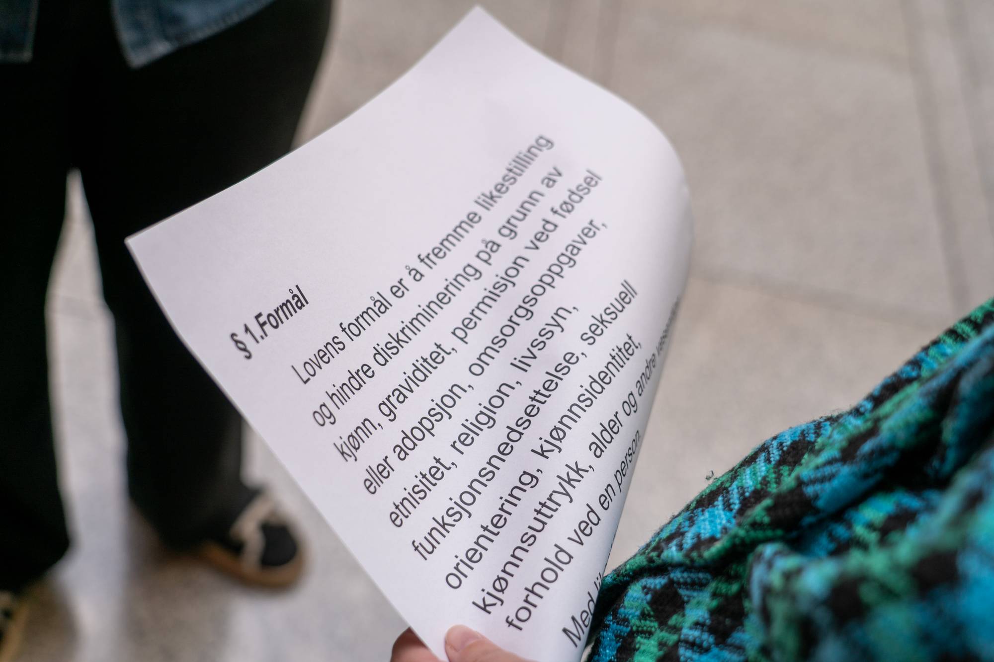 En person holder et dokument med tekst. Dokumentet inneholder paragrafen §1.Formål og tekst om likestilling og diskriminering. Bakgrunnen er uskarp, men man ser deler av en person og en grønn og blå rutete jakke. (Bildebeskrivelsen er laget av en KI-tjeneste)