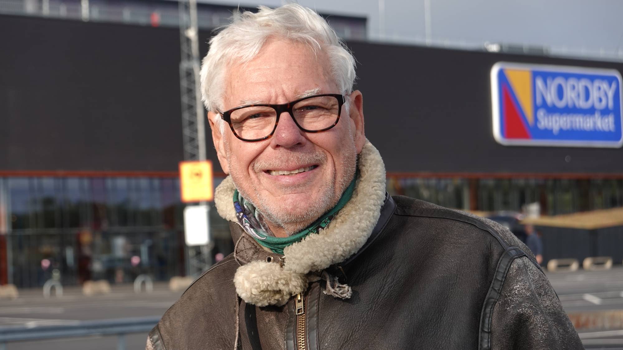 Eldre mann med briller og skinnjakke foran Nordby Supermarket. (Bildebeskrivelsen er laget av en KI-tjeneste)