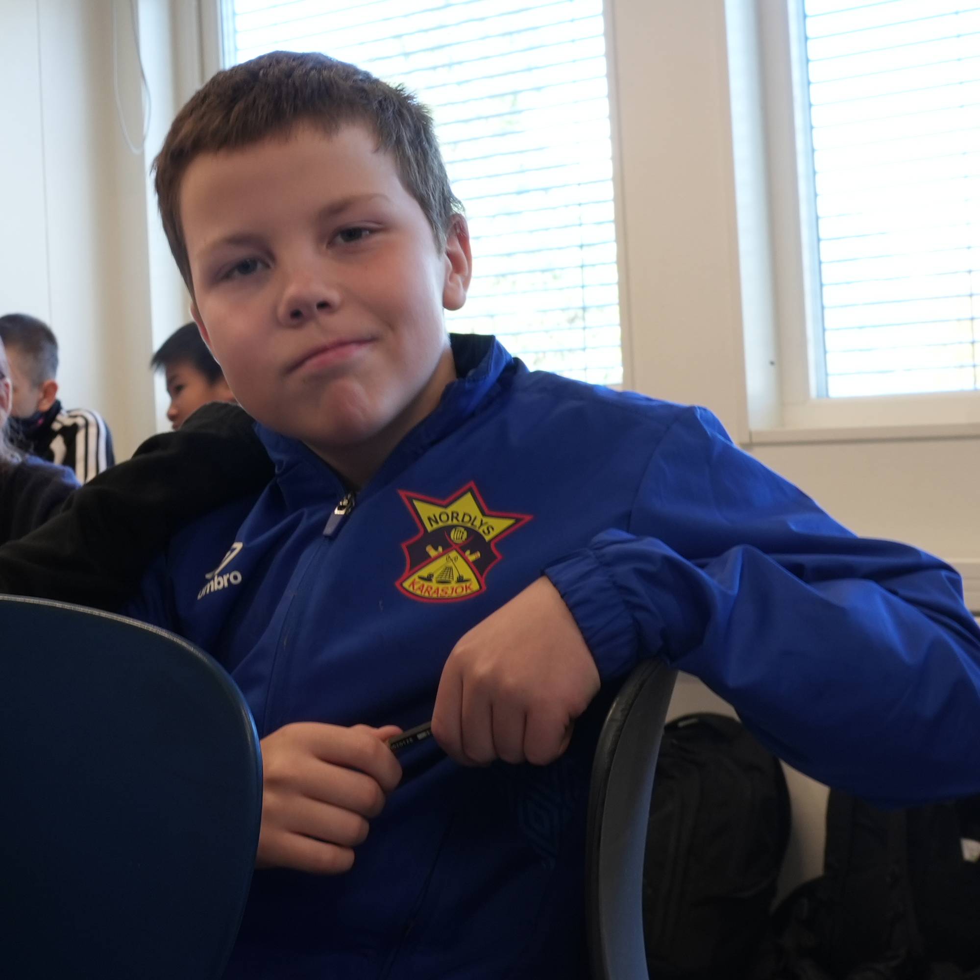 To gutter sitter ved en pult i et klasserom og ser mot kamera. Andre elever sitter i bakgrunnen. (Bildebeskrivelsen er laget av en KI-tjeneste)