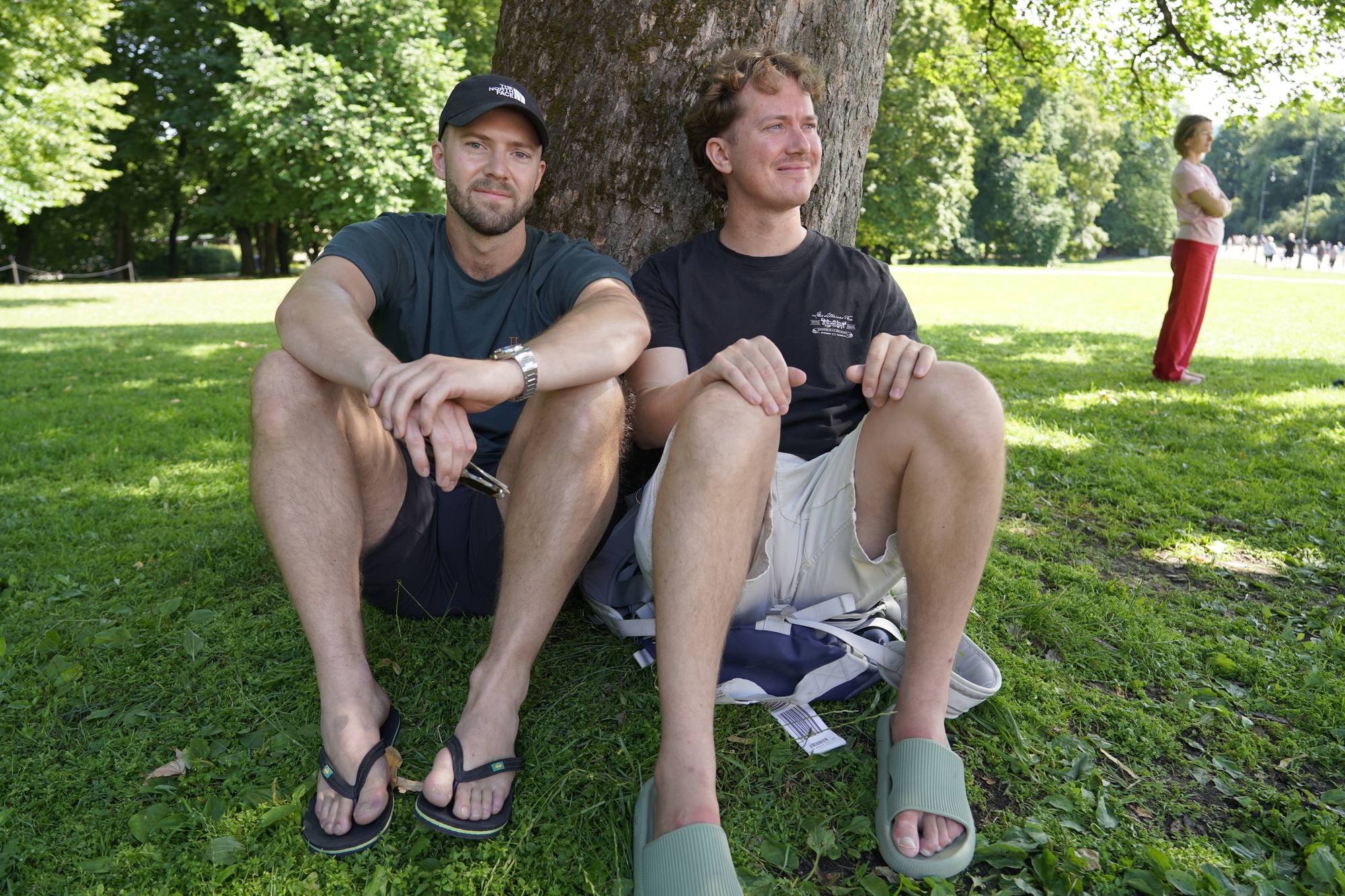 To menn sitter på gresset inntil et tre i en park. En kvinne står i bakgrunnen. (Bildebeskrivelsen er laget av en KI-tjeneste)