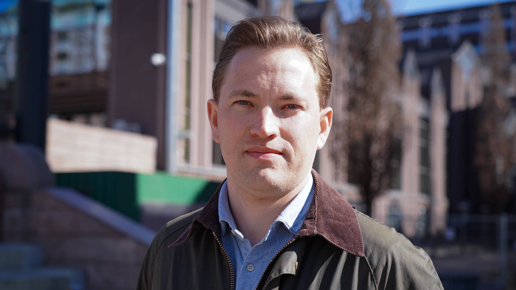 En mann med kortklipt hår og et lyseblått skjorte med krage, står foran en bygning. Han har en mørk jakke med brun krage. Mannen ser mot kamera. (Bildebeskrivelsen er laget av en KI-tjeneste)