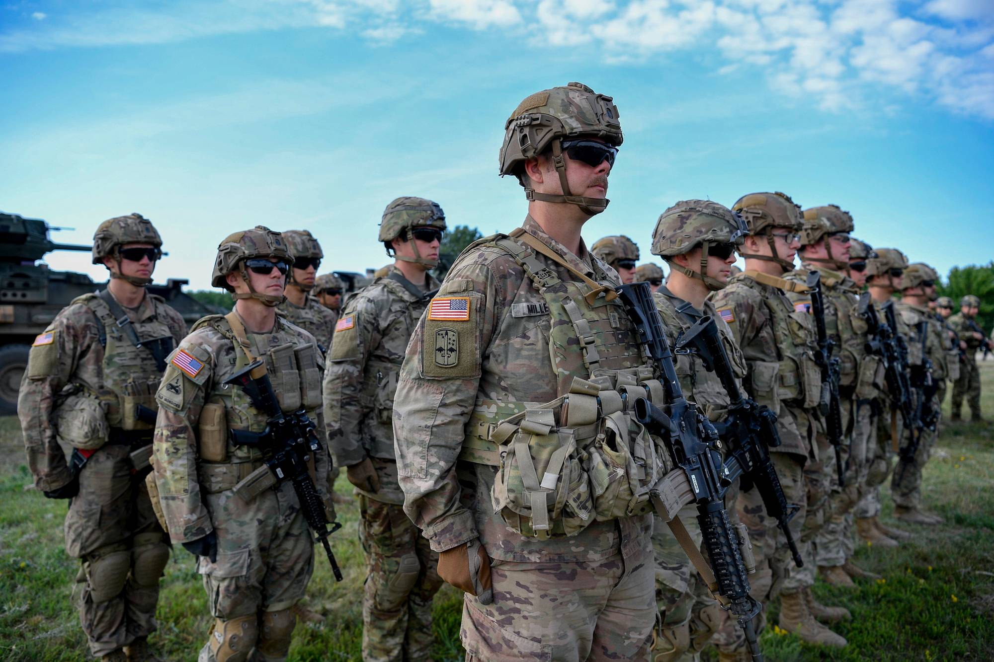 Soldater står på rekke i militæruniformer. De bærer hjelmer og solbriller, og har rifler ved siden av seg. Bak dem er det grønt gress og en blå himmel. Til venstre i bildet er en militærkjøretøy. (Bildebeskrivelsen er laget av en KI-tjeneste)