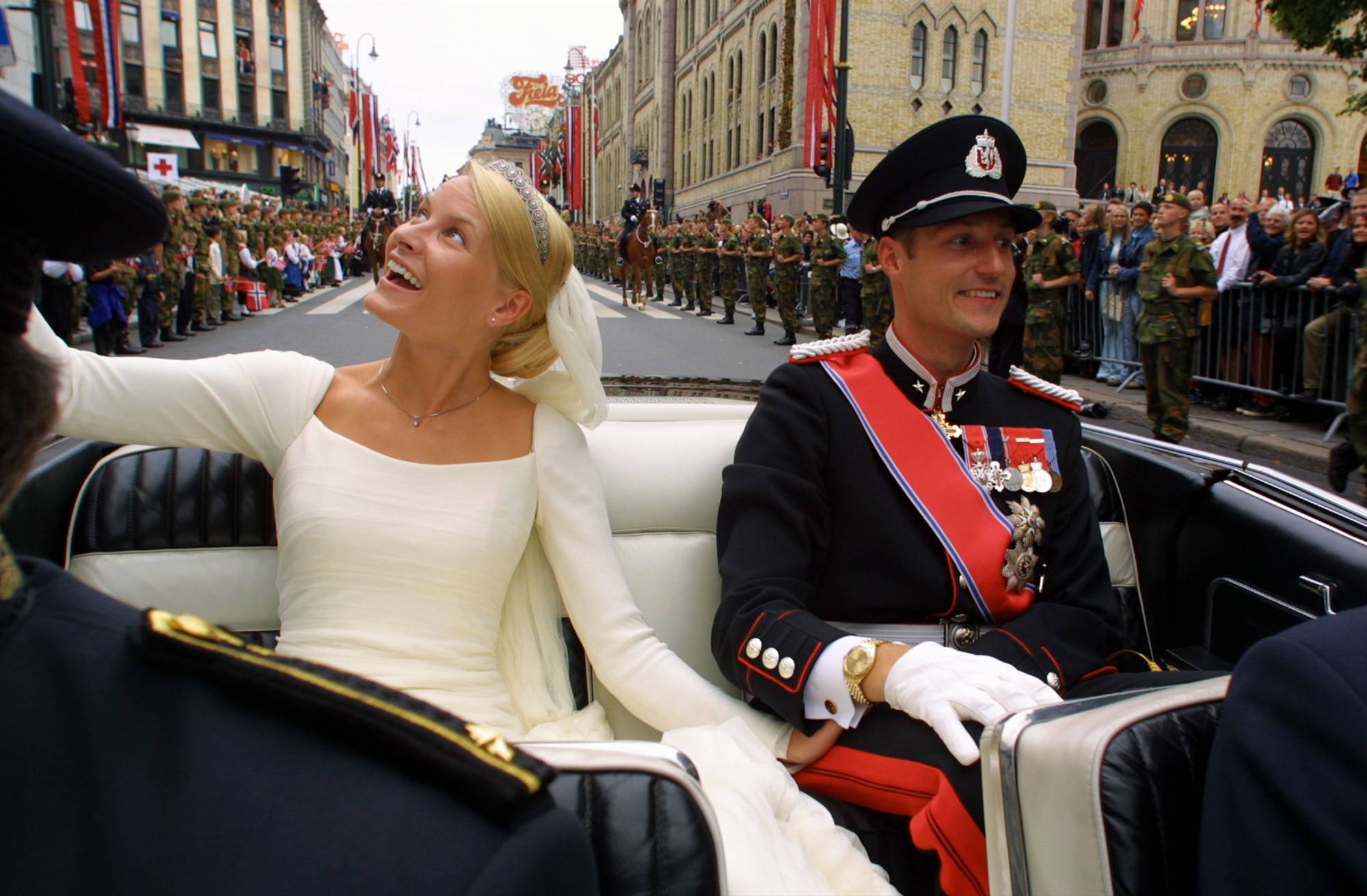 Kronprins Haakon og kronprinsesse Mette-Marit sitter i en åpen bil etter å ha blitt viet i Oslo domkirke og smiler og vinker til de mange som står langs Karl Johans gate opp mot Slottet