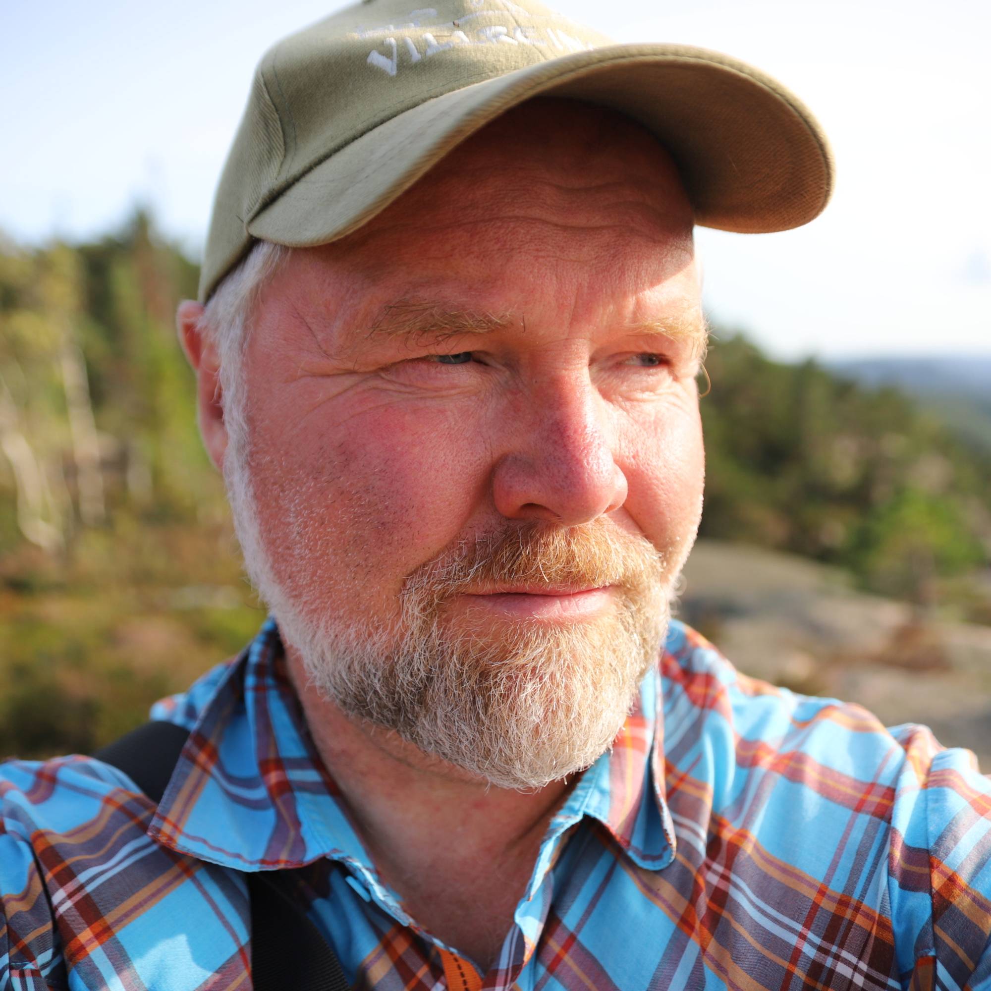 Mann med grått hår og skjegg med lysegrønn caps og blå- og rød-rutete skjorte ser utover i fjellnatur.