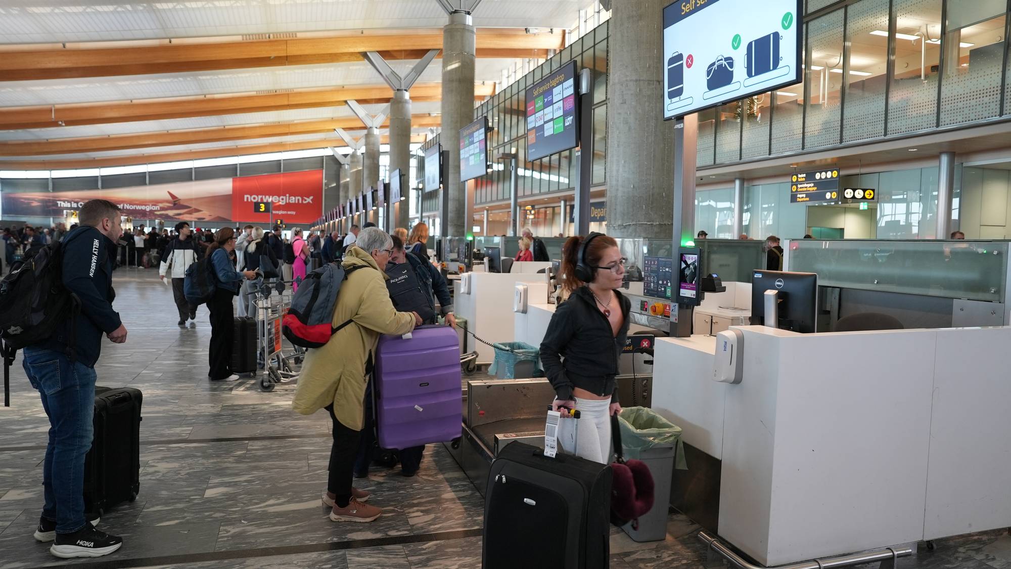 Reisende sjekker inn bagasje ved selvbetjente automater i en travel flyplasshall. (Bildebeskrivelsen er laget av en KI-tjeneste)