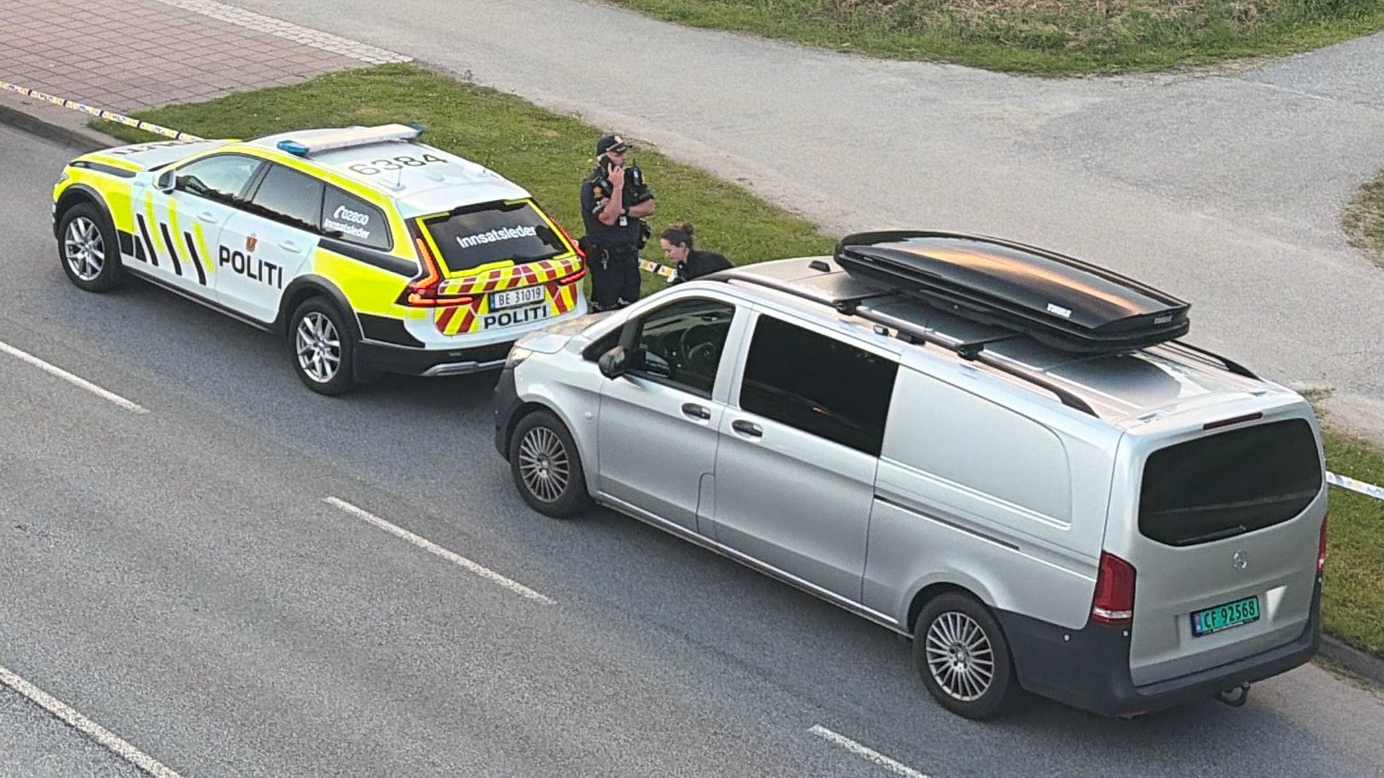 Politibetjenter og biler ved en sperret vei. Et område er markert med politisperrebånd. (Bildebeskrivelsen er laget av en KI-tjeneste)