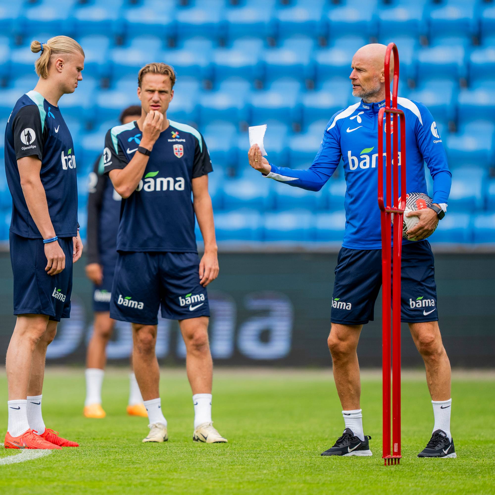 Trenere og spillere på en fotballbane. En trener holder et papir og en fotball, mens flere spillere står rundt ham. Bak dem ser man tomme tribuner. (Bildebeskrivelsen er laget av en KI-tjeneste)