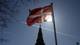 Tom Little / REUTERS,NTB Danmarks flagg vaier på en flaggstang mot en klarblå himmel. Solen skinner bak flagget. Nederst i bildet ses silhuetten av et tårn og bygningsmasse. (Bildebeskrivelsen er laget av en KI-tjeneste)