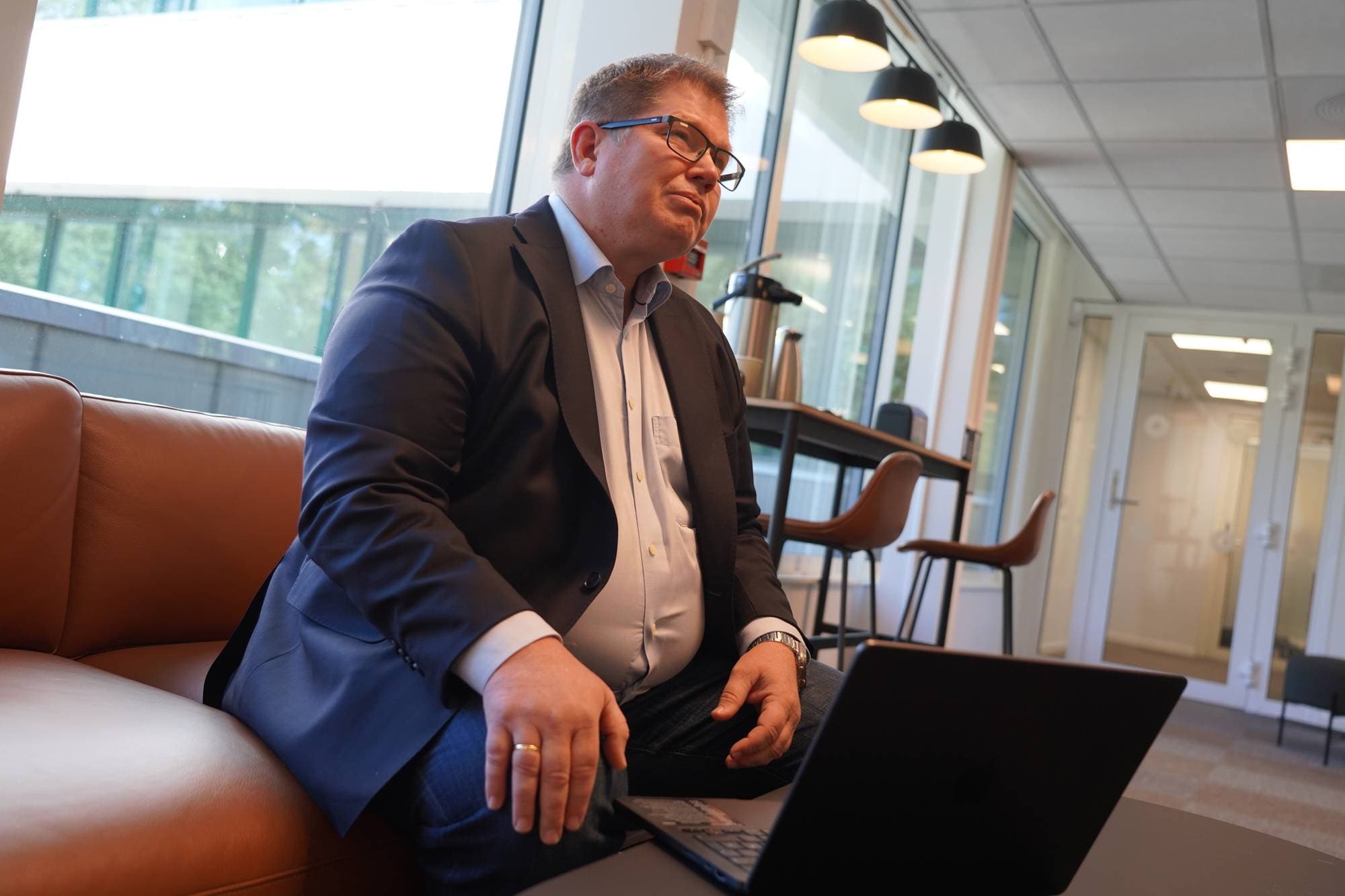 Mann i dressjakke sitter i en sofa med en bærbar datamaskin foran seg på et kontor. (Bildebeskrivelsen er laget av en KI-tjeneste)