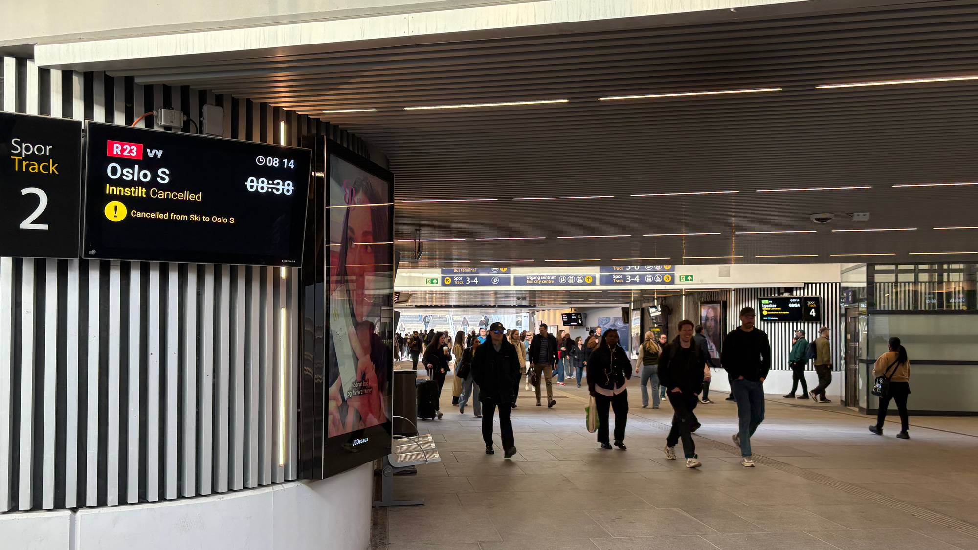 En skjerm viser at et tog til Oslo S er innstilt. Flere personer går gjennom en moderne togstasjon. (Bildebeskrivelsen er laget av en KI-tjeneste)