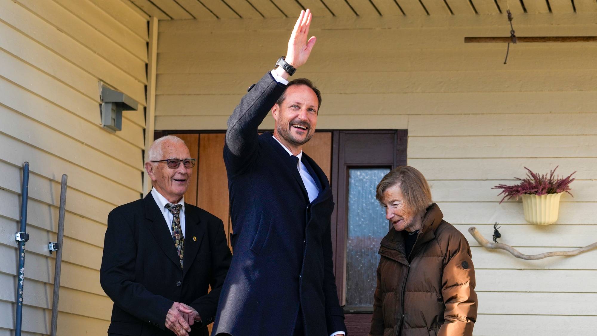 Kronprins Haakon vinker til folkemengden sammen med to eldre personer foran et hus. (Bildebeskrivelsen er laget av en KI-tjeneste)