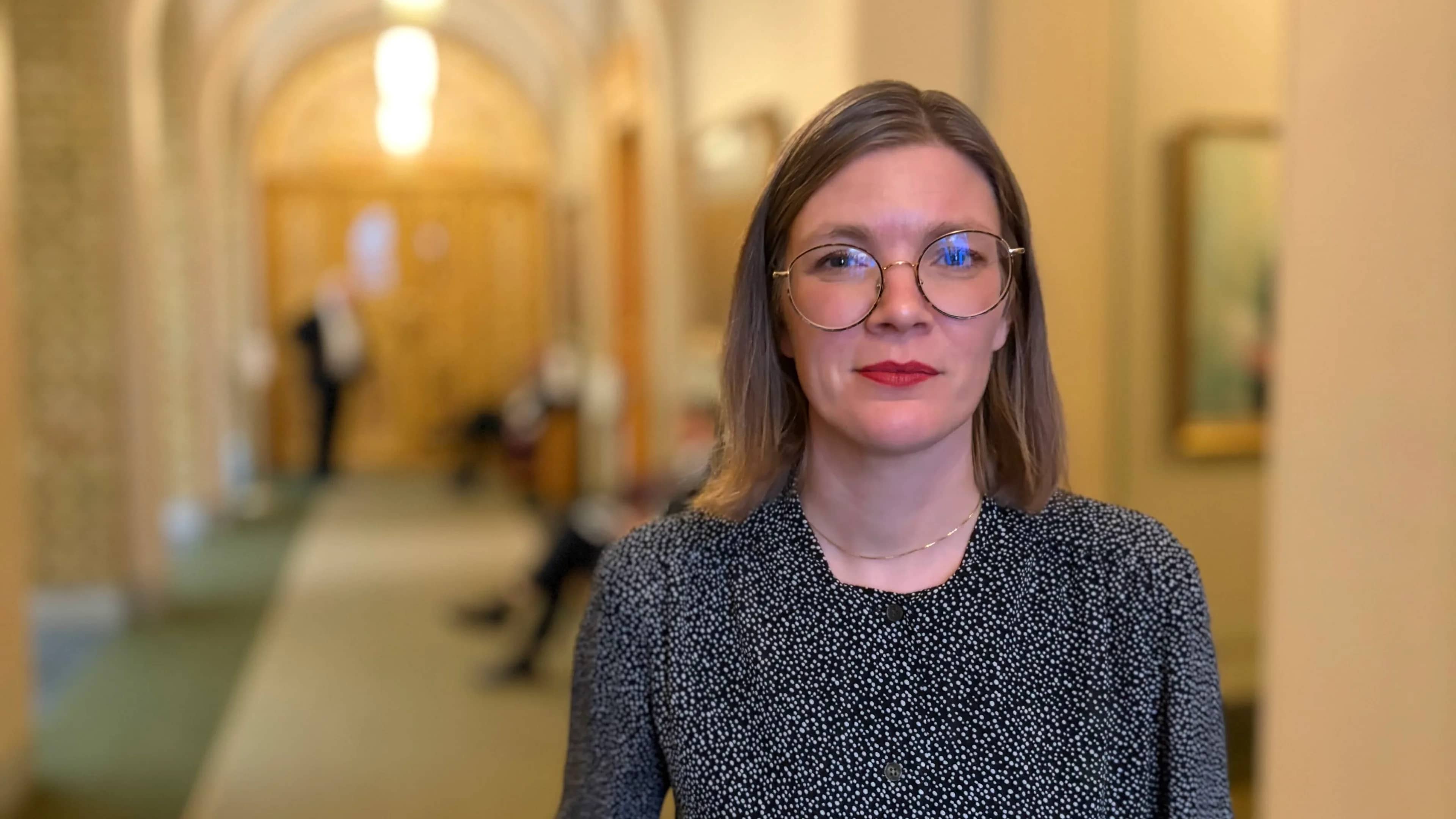 Marie Sneve Martinussen står i gangen på Stortinget. Hun har skulderlangt, stritt hår og briller. Hun står alvorlig med hendene i lommen.
