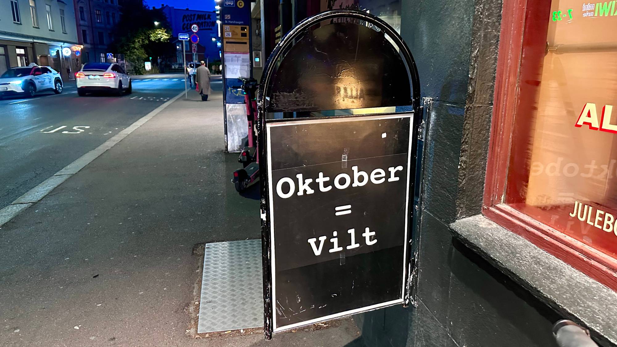 Skilt på fortau med teksten Oktober og Vilt, plassert utenfor et utested i en bygate om kvelden.