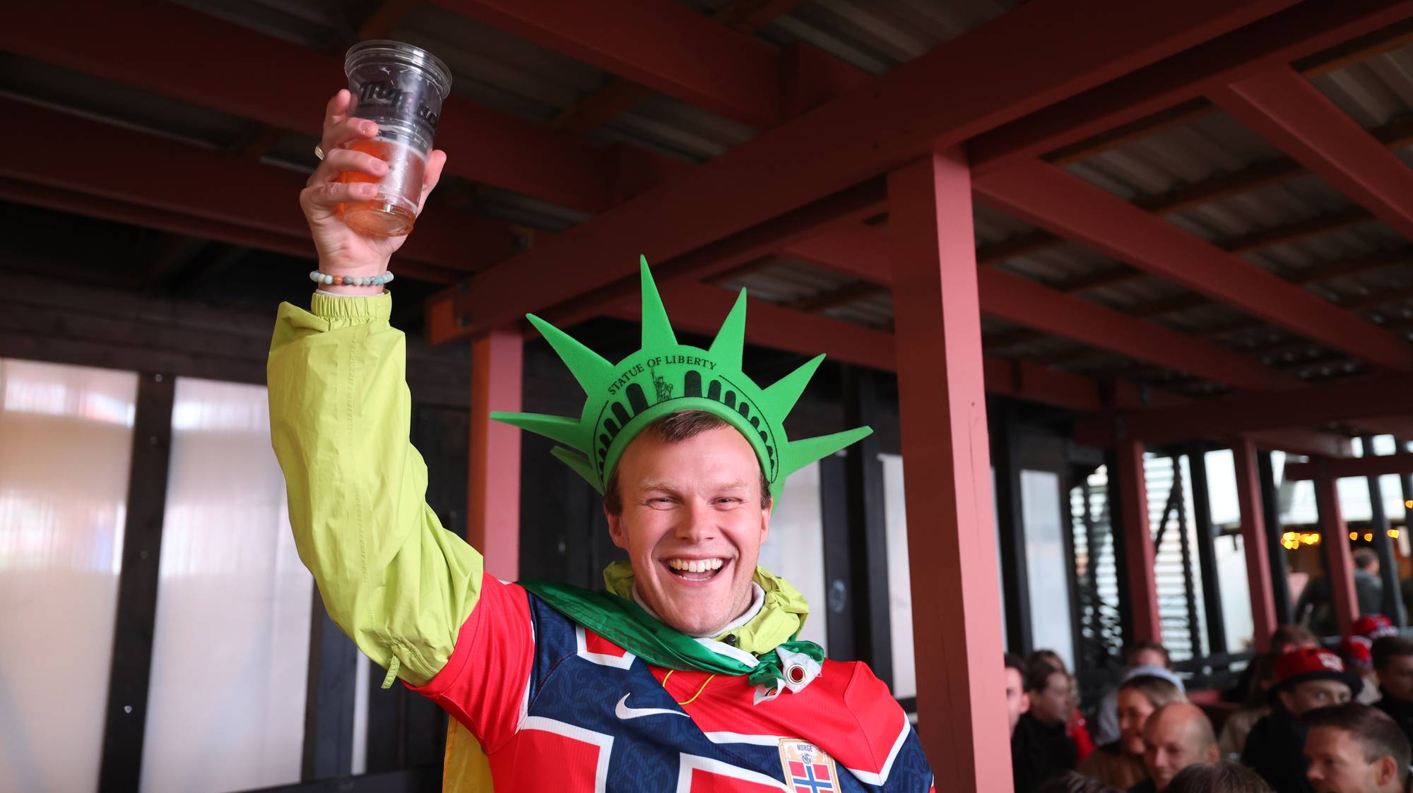 Person med Frihetsgudinnen-hatt og norsk landslagsdrakt hever et plastglass i et lokale. (Bildebeskrivelsen er laget av en KI-tjeneste)