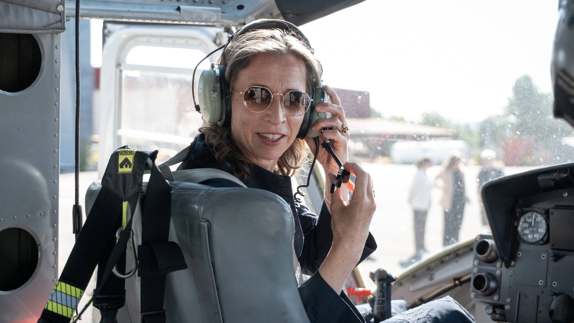 Kvinne sitter i et helikopter med hodesett og solbriller. Bakgrunnen viser et uteområde. (Bildebeskrivelsen er laget av en KI-tjeneste)