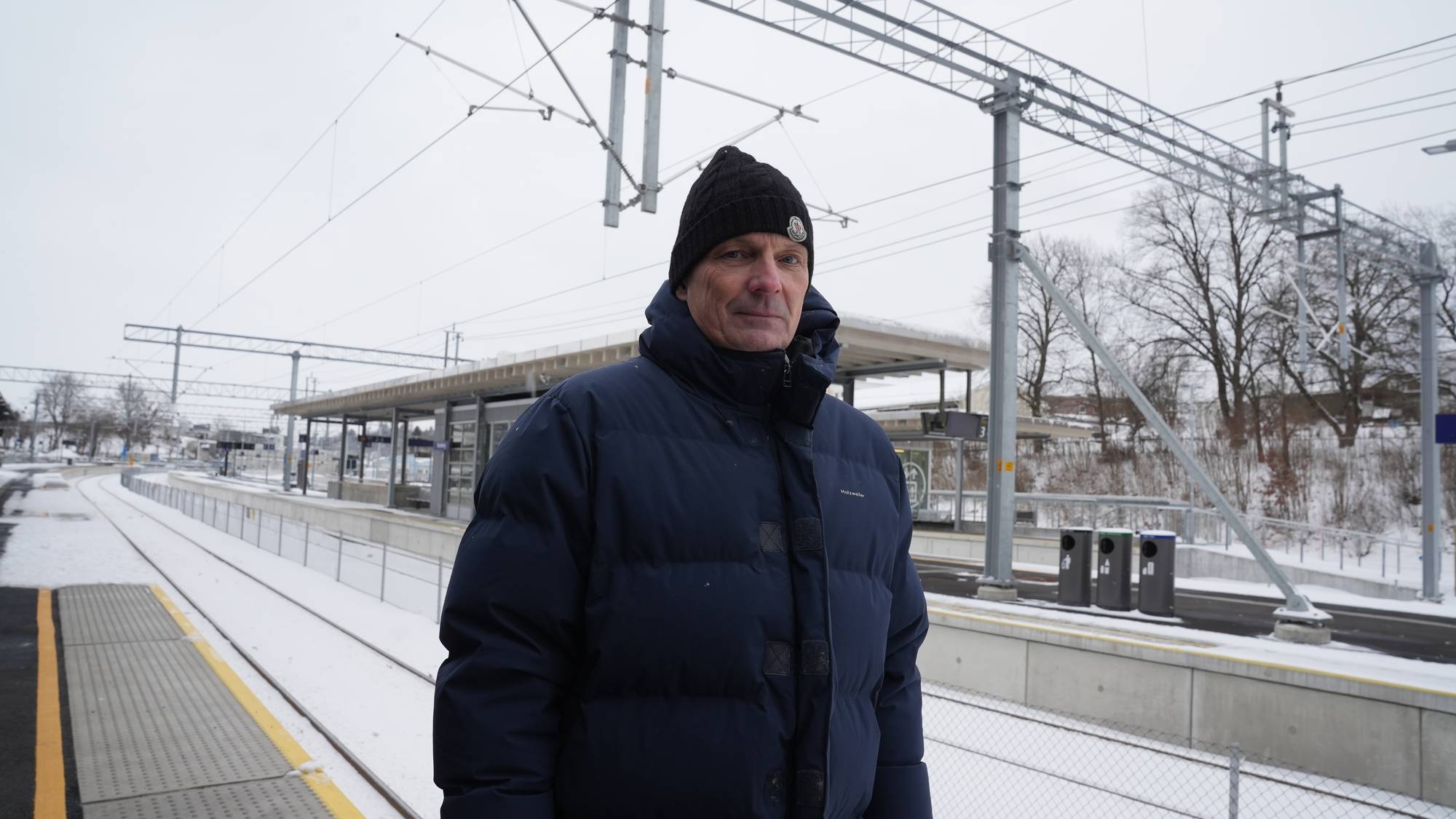 En person står på en snødekt plattform ved en jernbanestasjon. Personen er iført en blå jakke og lue. I bakgrunnen sees en moderne stasjonsbygning samt jernbanelinjene som strekker seg bortover. Omgivelsene er preget av vintervær med snø og grå himmel. (Bildebeskrivelsen er laget av en KI-tjeneste)