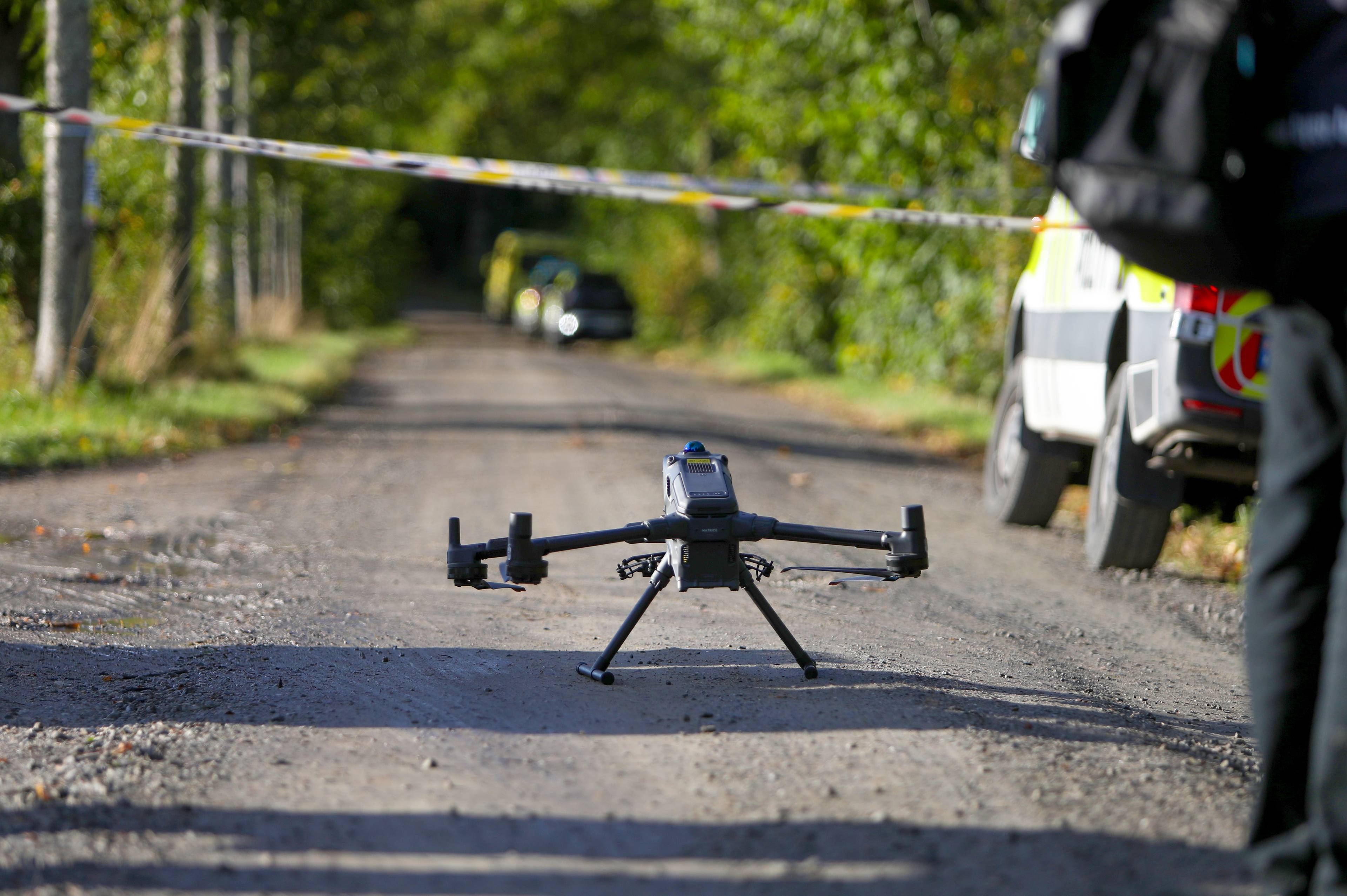 En drone står på en grusvei ved et politisperret område med en politibil i bakgrunnen. (Bildebeskrivelsen er laget av en KI-tjeneste)