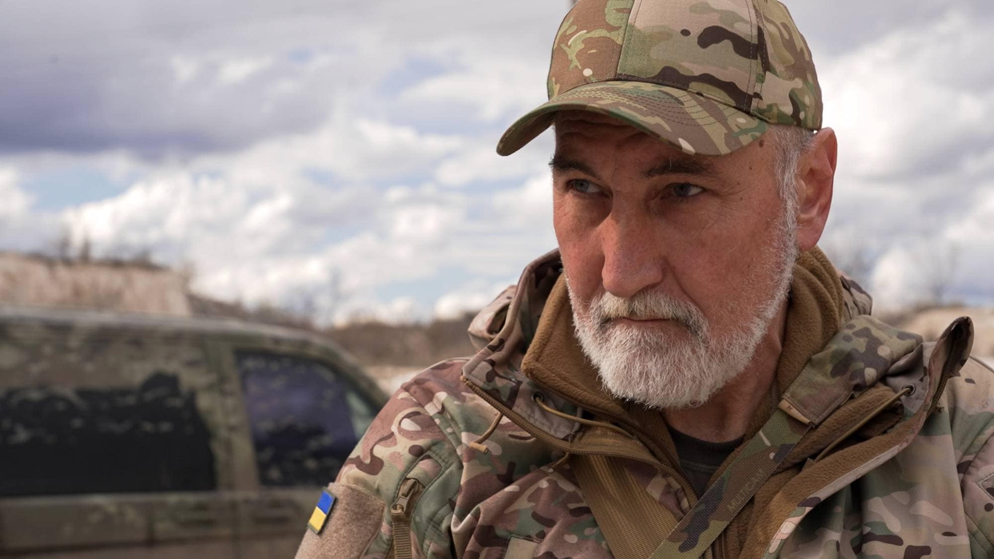En mann i kamuflasjeklær og caps ser til siden. En militærbil står i bakgrunnen. Mannen har et grått skjegg. Det er et ukrainsk flagg på vesten hans. (Bildebeskrivelsen er laget av en KI-tjeneste)