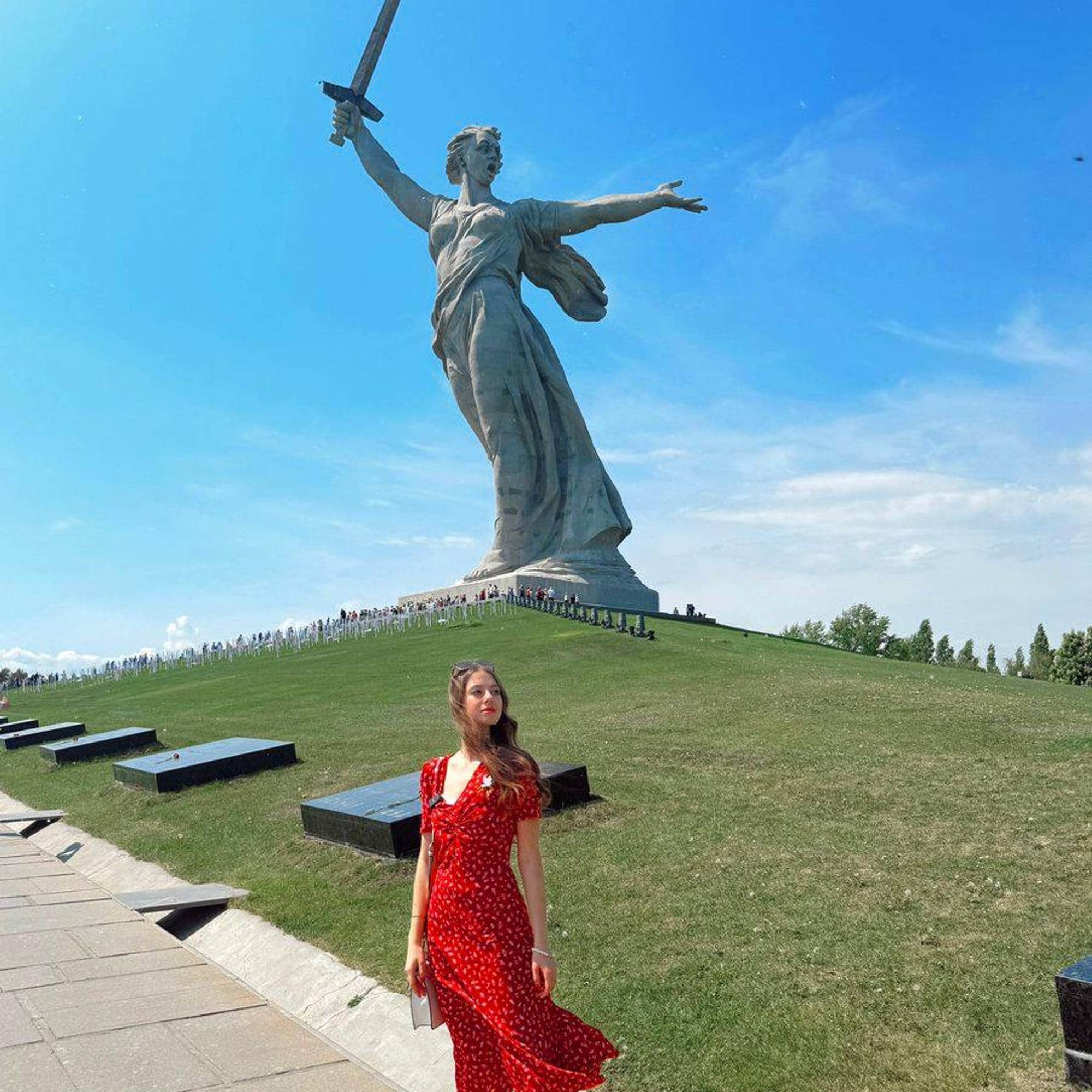 Kvinne står i en rød kjle foran en statue med en kvinne som holder et sverd
