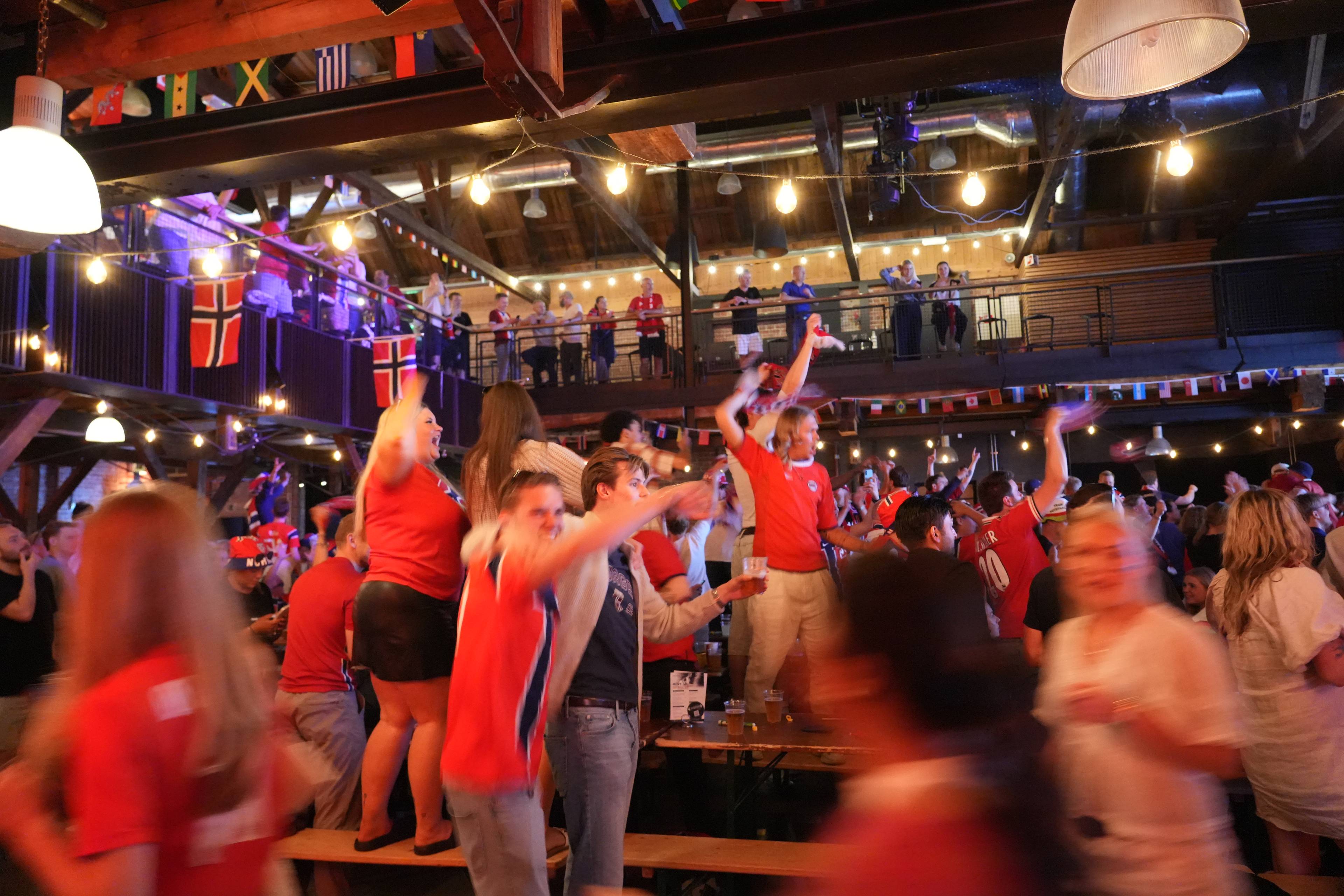 En folkemengde med norske flagg og trøyer feirer inne i et lokale. Flere personer danser og jubler. I bakgrunnen er det en scene med flere personer som ser ned på folket i etasjen under. Det er pyntet med flagg fra ulike nasjoner.